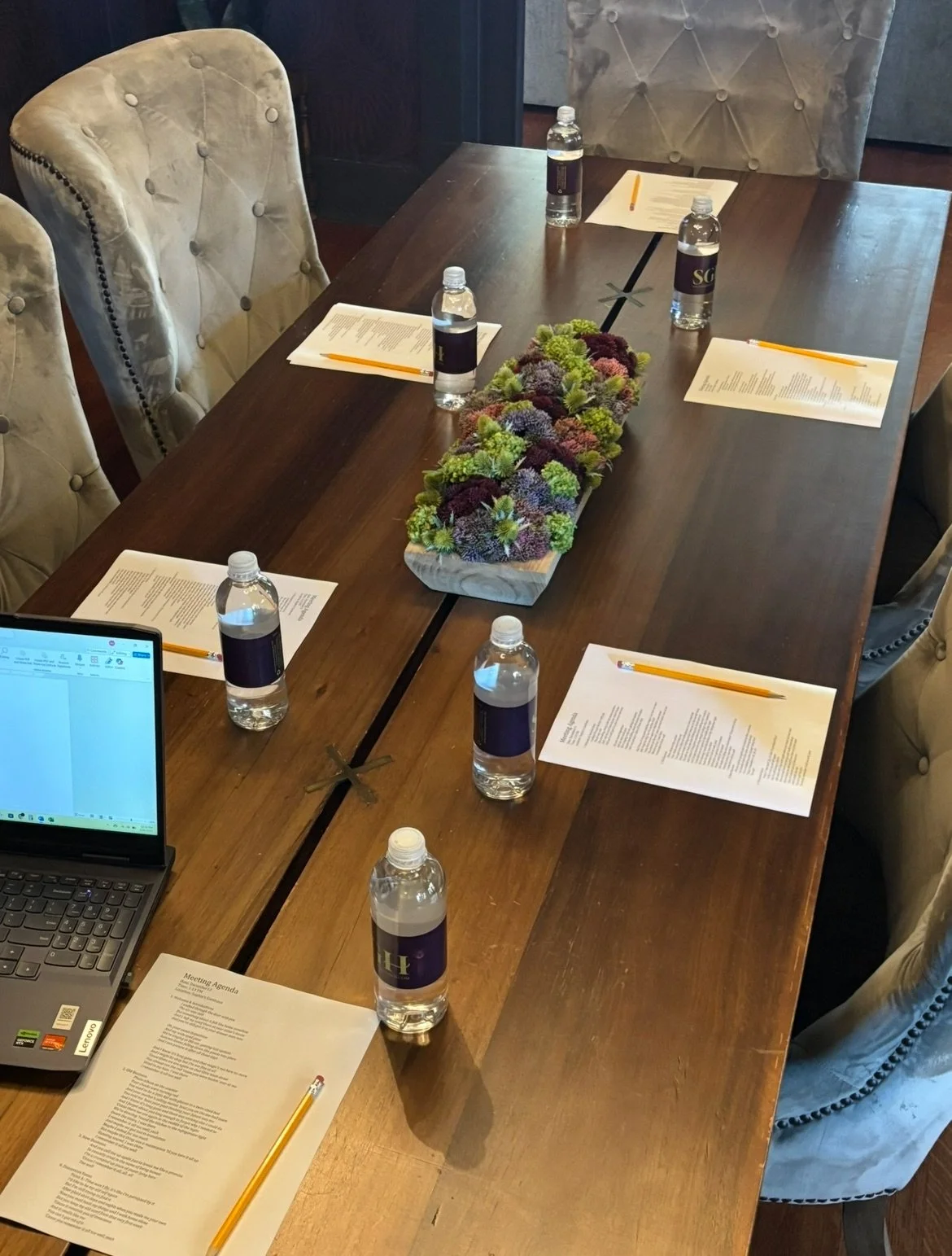 A conference table set for a meeting with six bottles of water, six printed agendas, and a laptop. There is a rectangular floral centerpiece with purple, green, and white flowers.