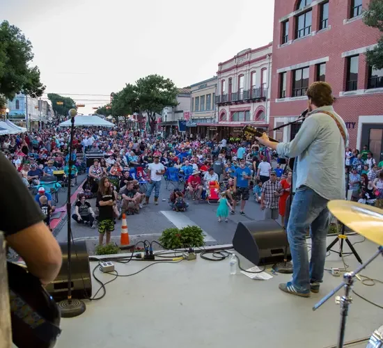 Musician performing on stage with a guitar, overlooking a lively street festival filled with people sitting and walking, surrounded by historic buildings.