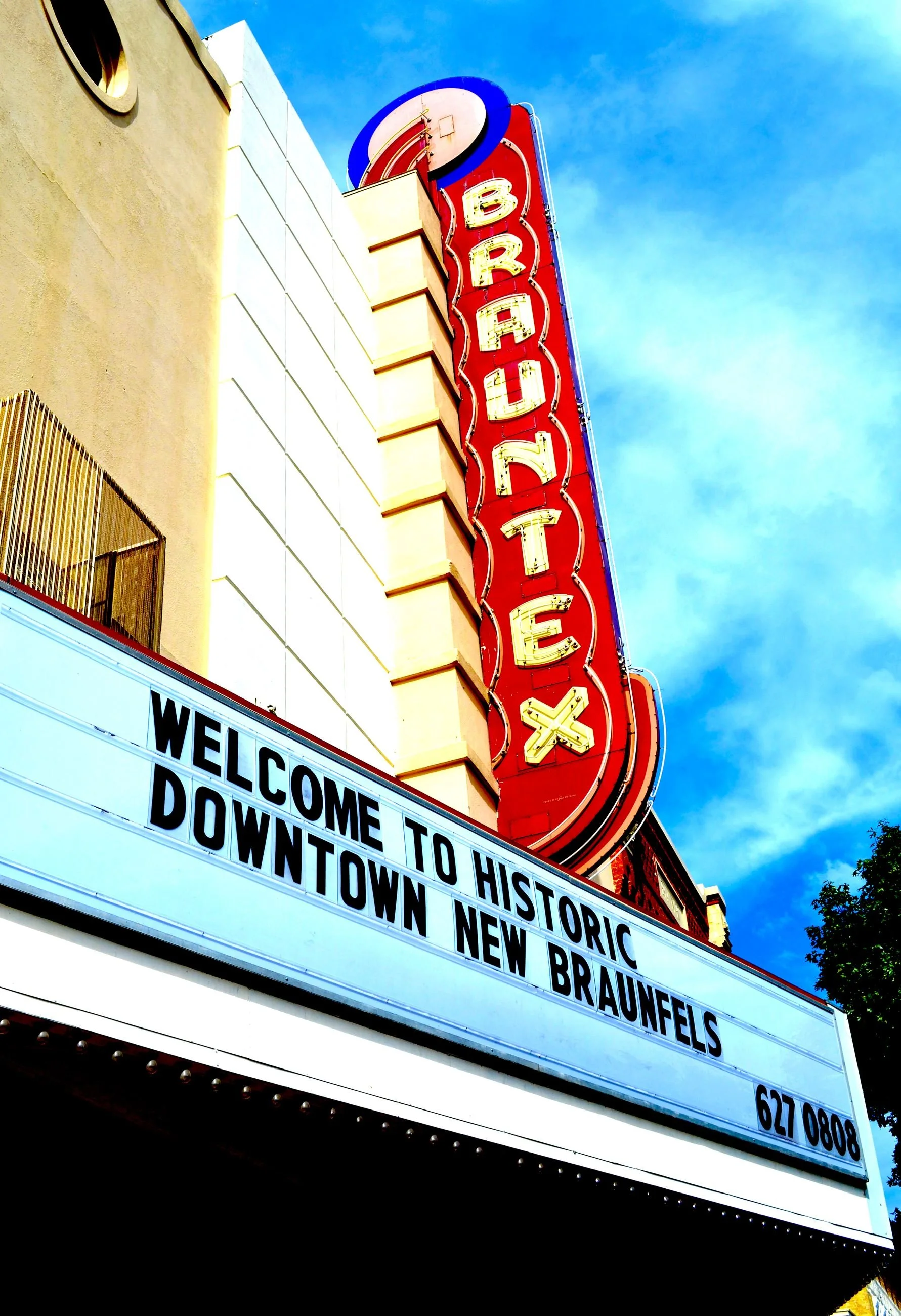 The image features a vintage-style vertical sign for Brauntelex, and a marquee board with a welcome message reading, 'Welcome toHistoric Downtown New Braunfels,' against a blue sky with some clouds.