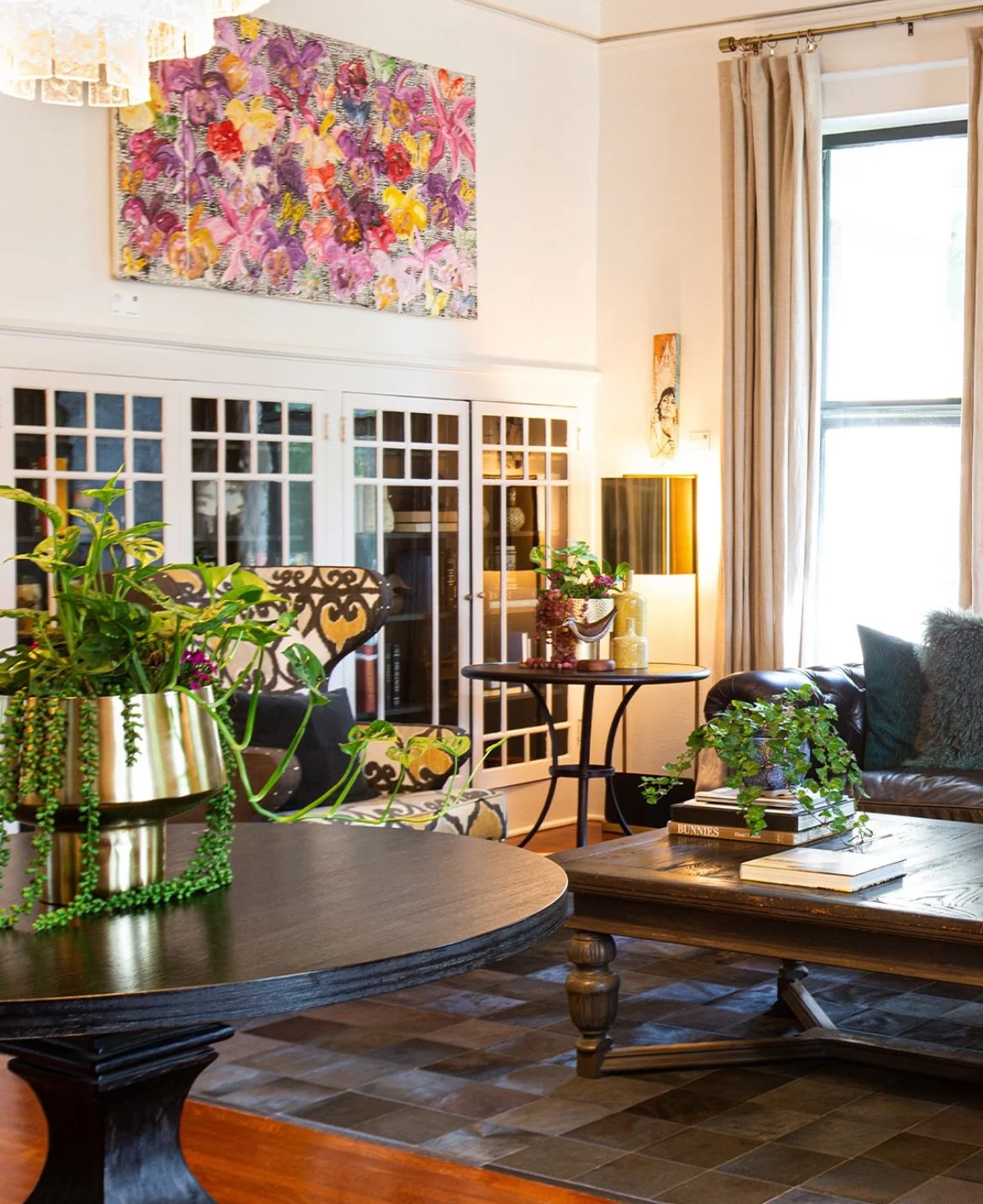 Living room with a colorful floral painting on the wall, a round black wooden coffee table with books and greenery, a patterned armchair, a brown leather sofa with pillows, a black side table with a plant and decorative items, and large windows with beige curtains letting in natural light.