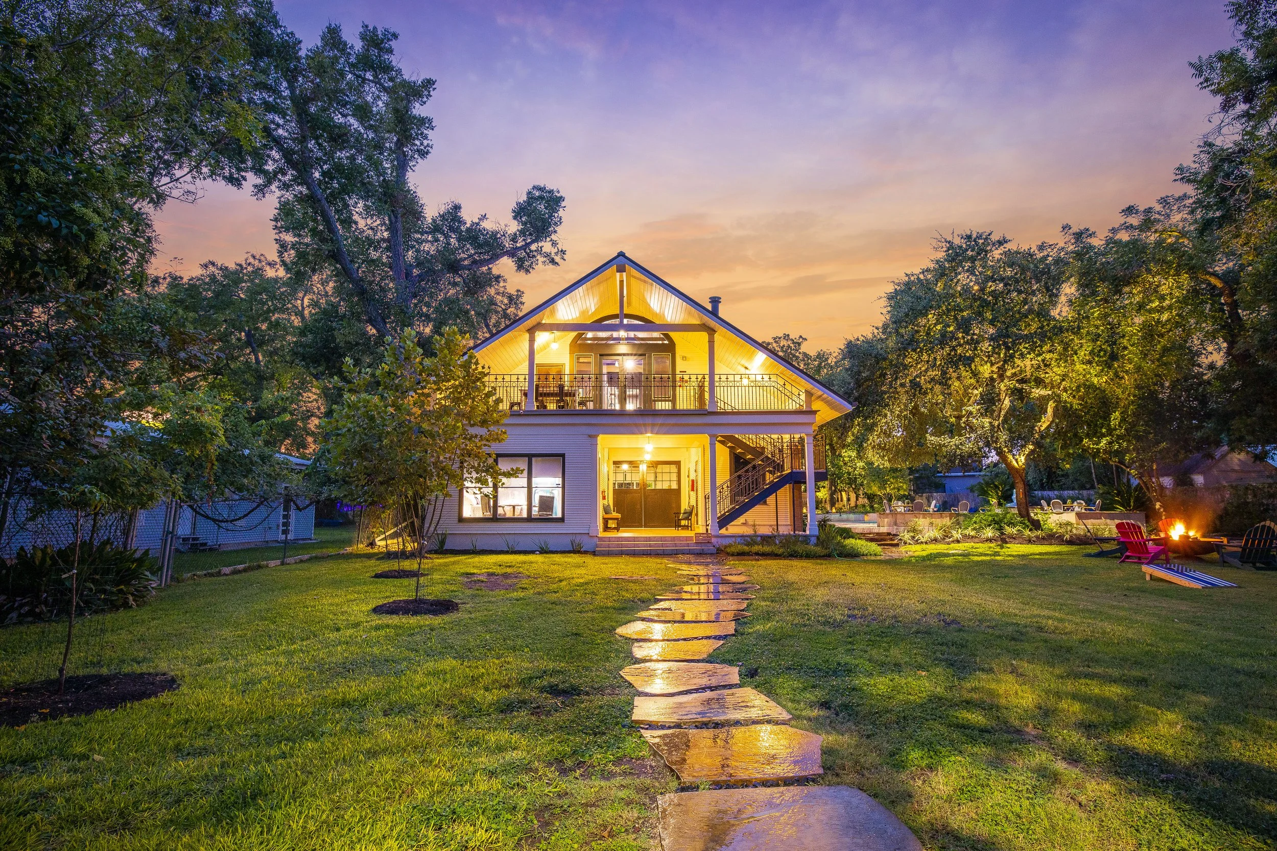 A two-story house illuminated at dusk with a lit-up porch and balcony, surrounded by a lush green yard with a stone pathway leading to the front door, tall trees on either side, and a firepit with chairs on the right.