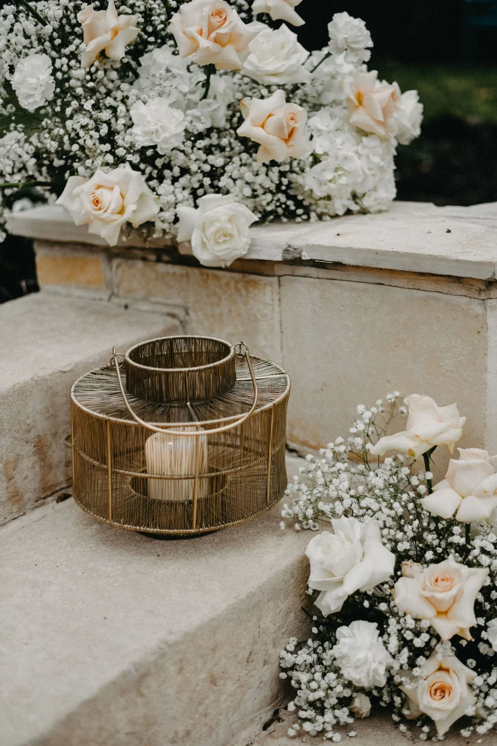 A floral arrangement with white roses and baby's breath on a stone ledge, accompanied by a decorative candle holder with a lit candle, against a dark background.