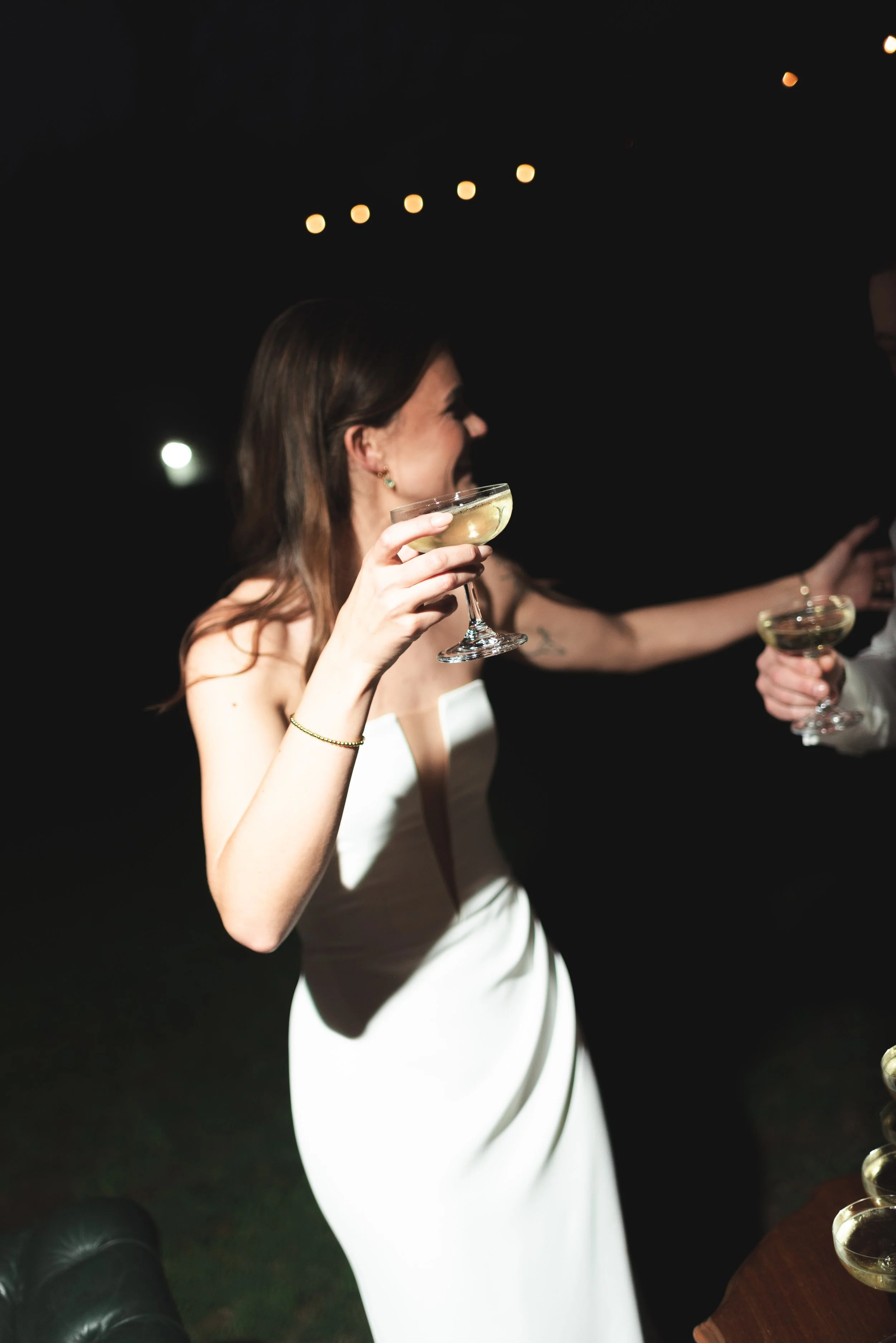 A woman in a white dress holding a champagne glass, smiling and toasting at a celebration or party.