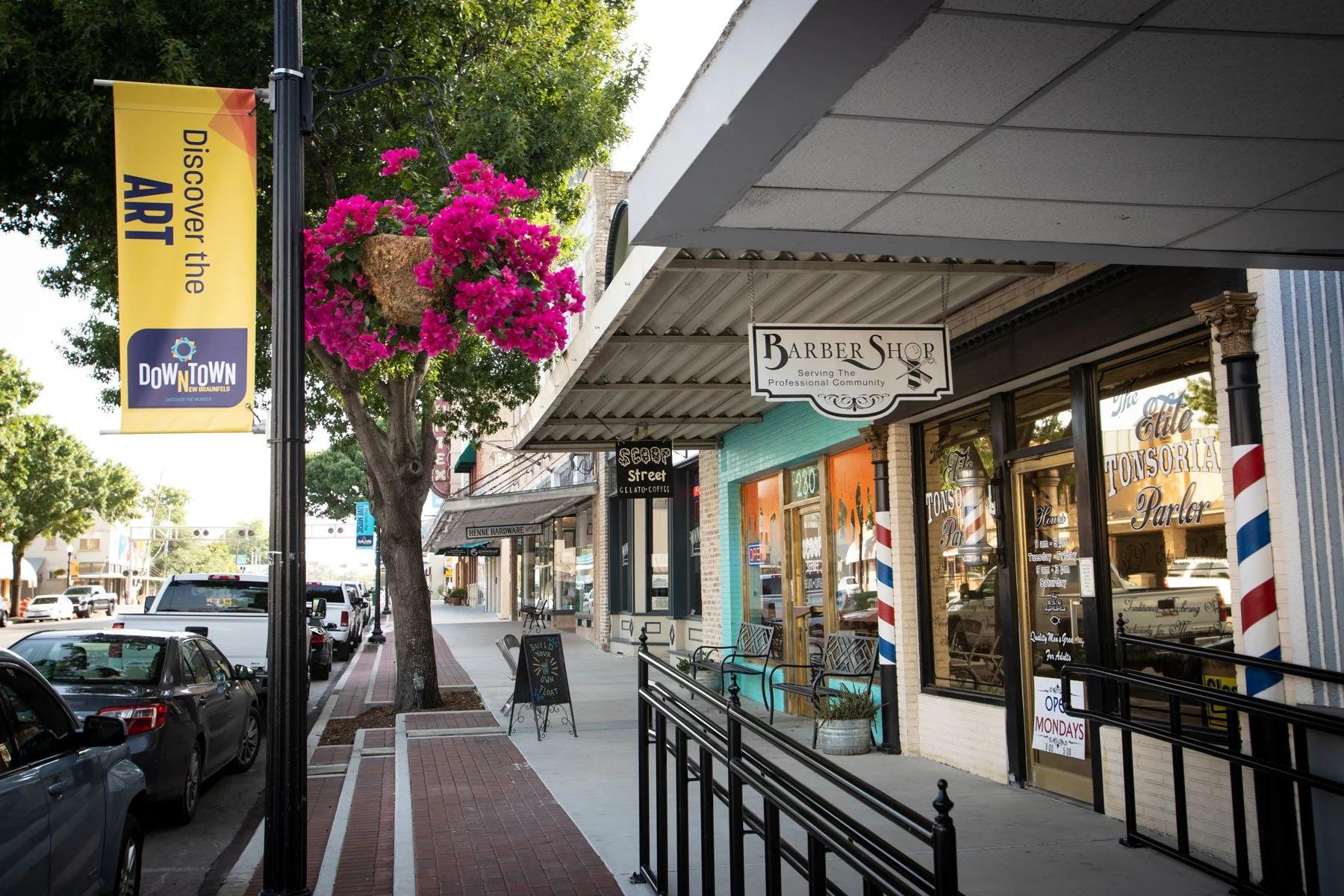 Downtown street with storefronts, parked cars, a flowering tree, and a yellow banner that reads "Discover the ART."