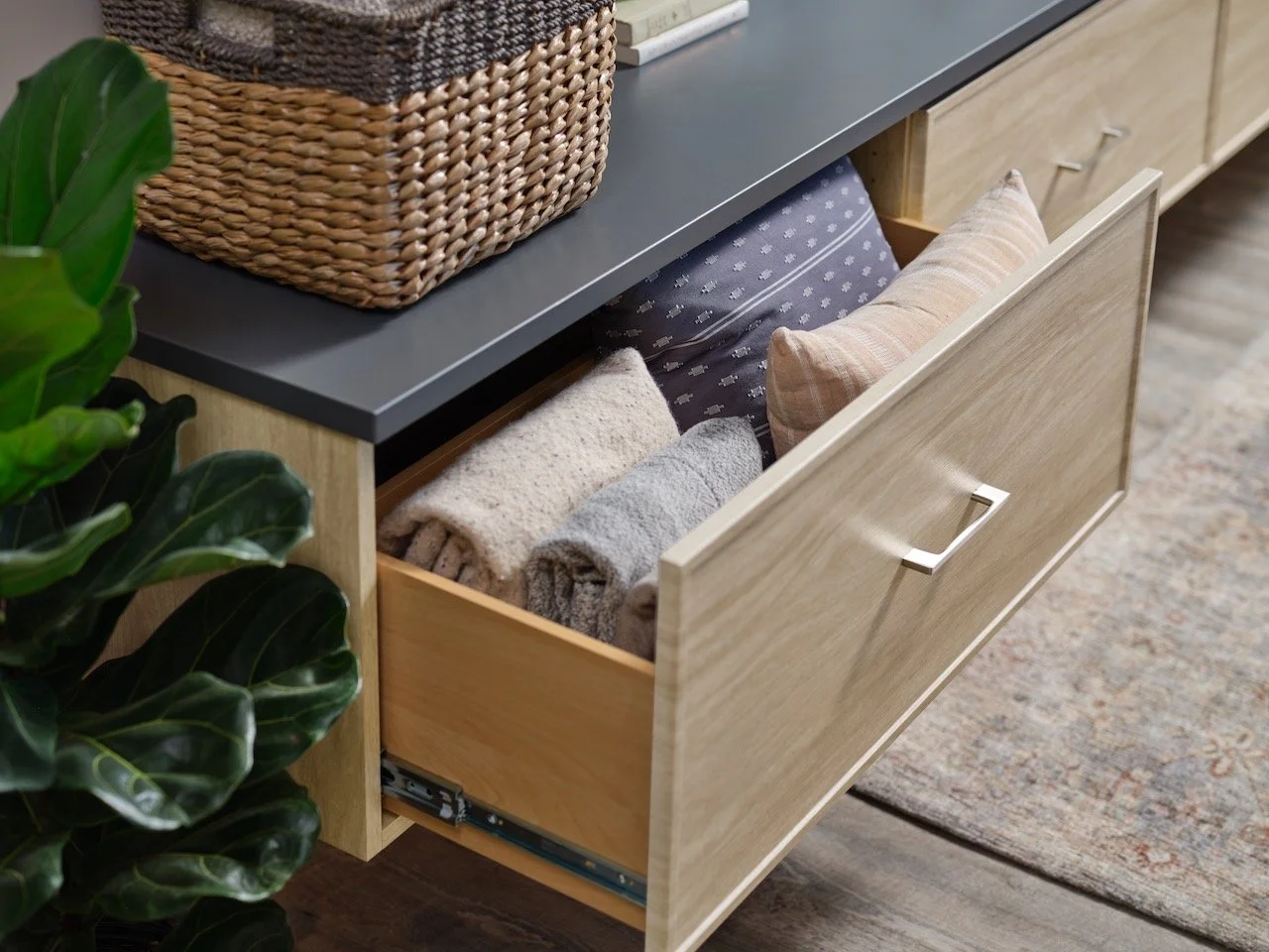 Open drawer in a wooden cabinet filled with folded blankets and a pillow, next to a green leafy plant and a woven basket on a black countertop.