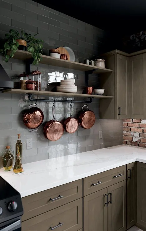 Kitchen with gray cabinets, white marble countertop, open wooden shelves holding dishes and jars, copper pots hanging, gray subway tile backsplash, brick wall in corner, small potted plant, and glass bottles on the counter.
