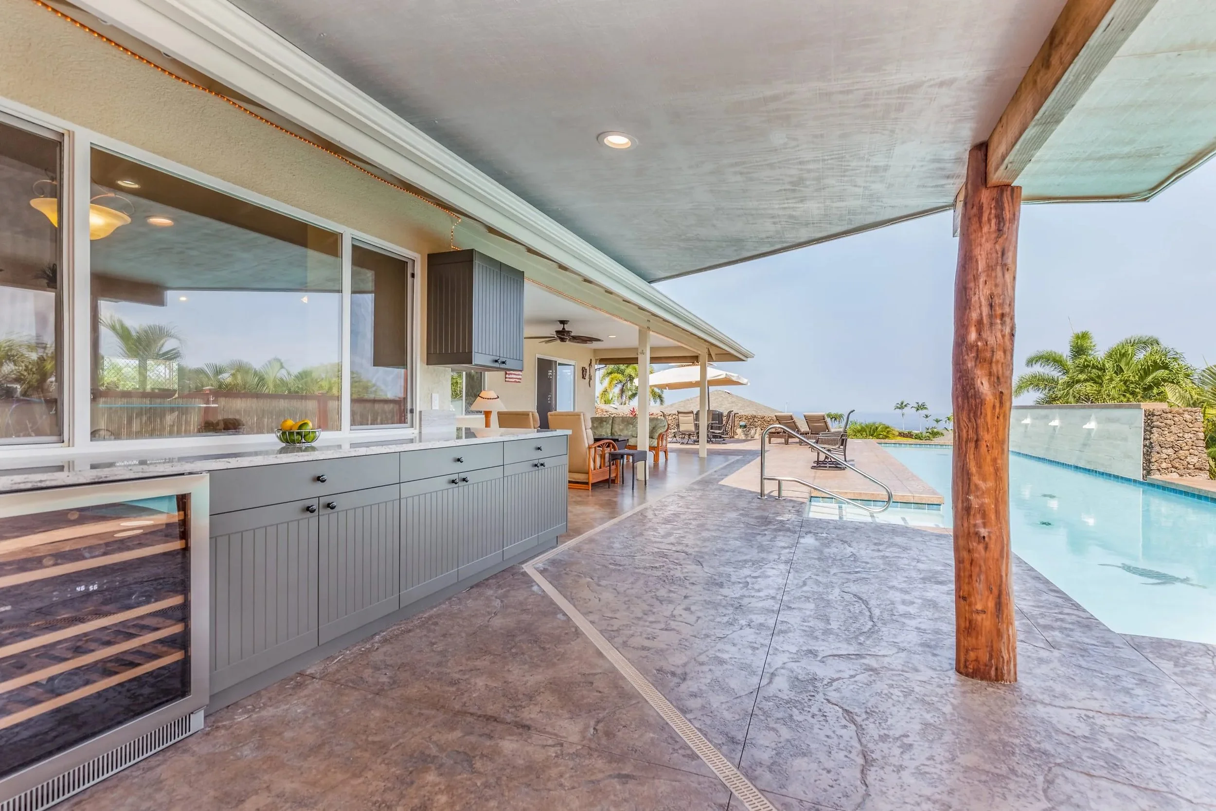 Outdoor covered pool area with lounge chairs, tables under umbrellas, and a kitchen or bar with grey cabinets and a wine cooler, with scenic tropical landscape in the background.