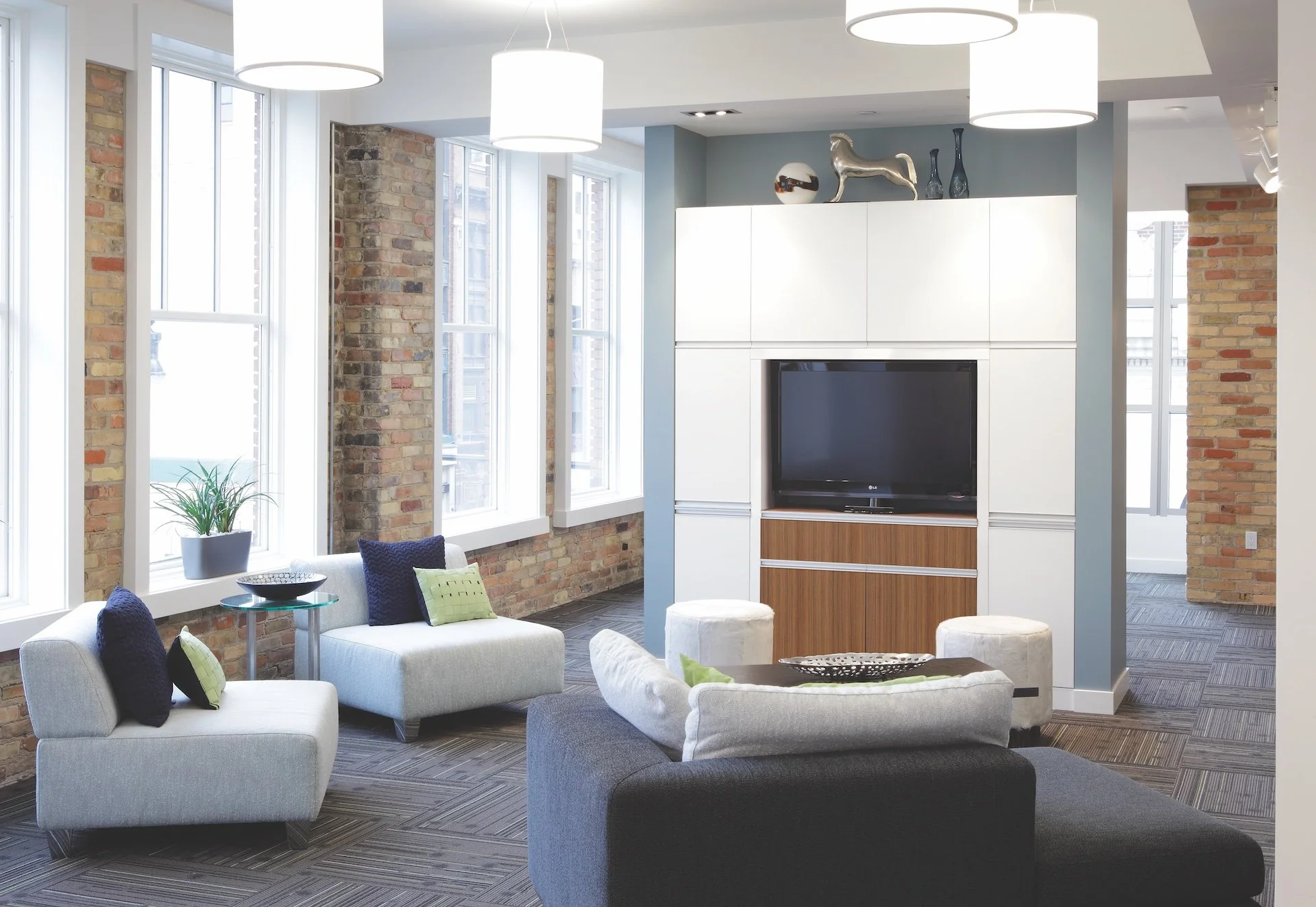 Modern living room with exposed brick walls, large windows, contemporary white and gray seating, colorful accent pillows, a flat-screen TV, and decorative items on a built-in wall unit.