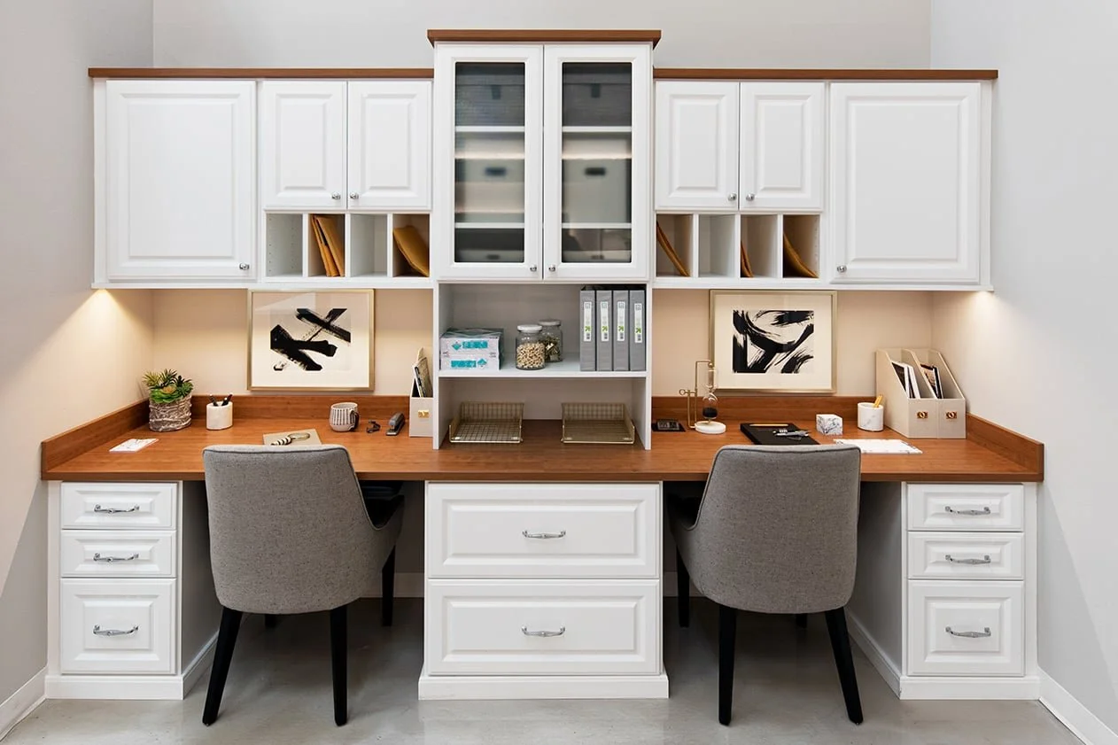 Modern office workspace with shared two seat wooden desk and white storage cabinets, two gray chairs, decorative plants, framed abstract art, office supplies, and shelving with binders and jars.