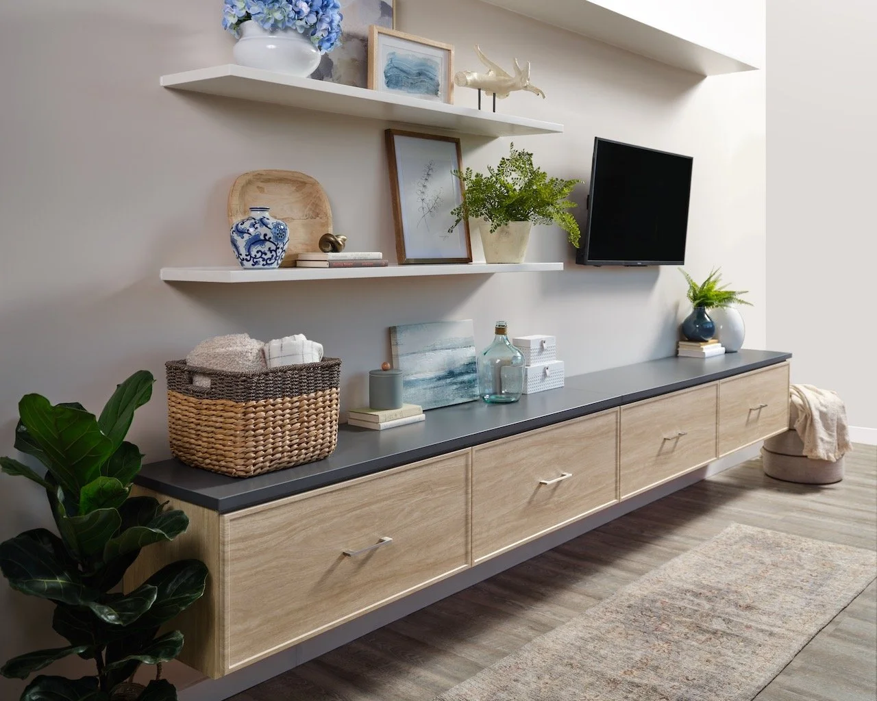 Modern living room with a light wood entertainment console, decorative books and vases, potted plants, artwork, and floating shelves with decorative items and a flat-screen TV.