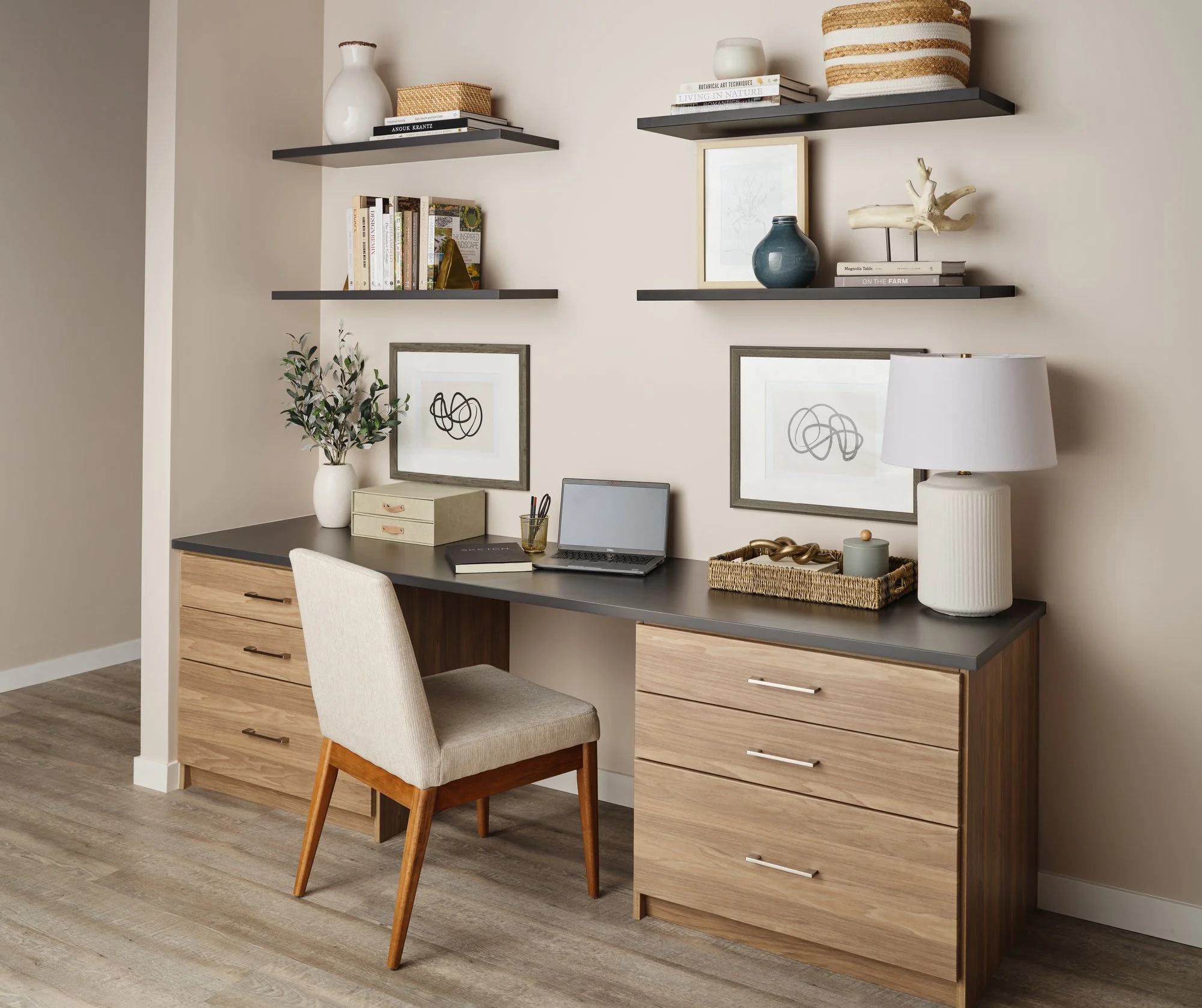 Home office desk with a beige upholstered chair, a white vase with greenery, books, framed abstract art, a laptop, and decorative items. Shelves above with more books, vases, and decorative objects, set against a plain light-colored wall.