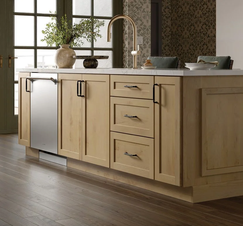 Kitchen with light wood cabinets, a marble countertop, and a brass faucet. Decor includes a large vase with green plants, a small dark stone, and a few dishes on the countertop. Large windows provide natural light to the space.