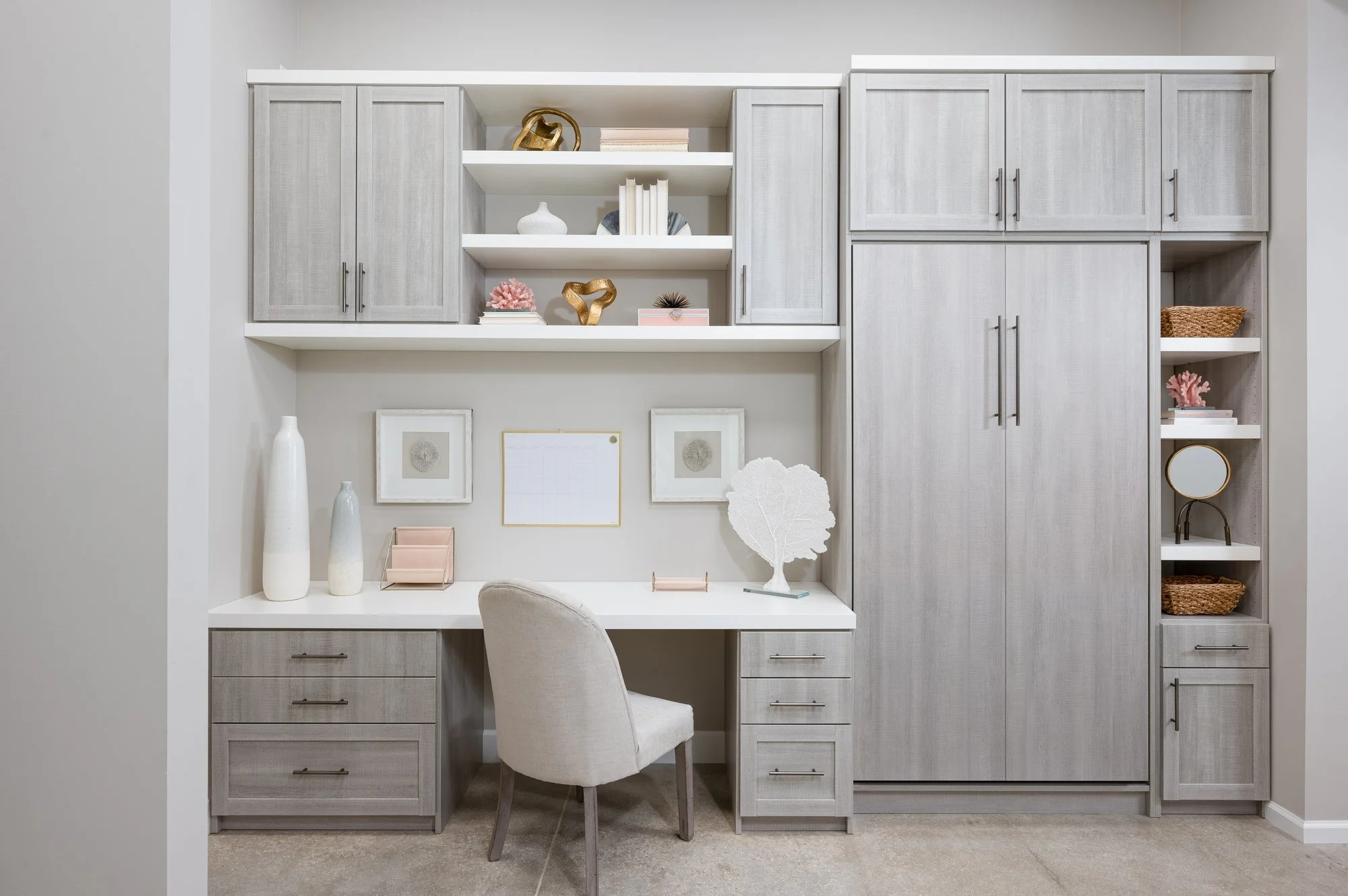 Home office with light gray built-in shelves, cabinets with a hidden murphy bed, and a white desk. Decor includes vases, books, art, and decorative objects, with a beige chair in front of the desk.