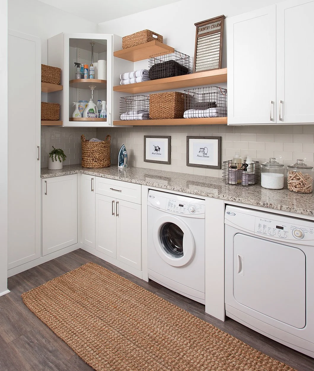Laundry room with white cabinets, washer and dryer, granite countertop, woven baskets, shelves with towels, laundry supplies, and framed pictures on the wall.