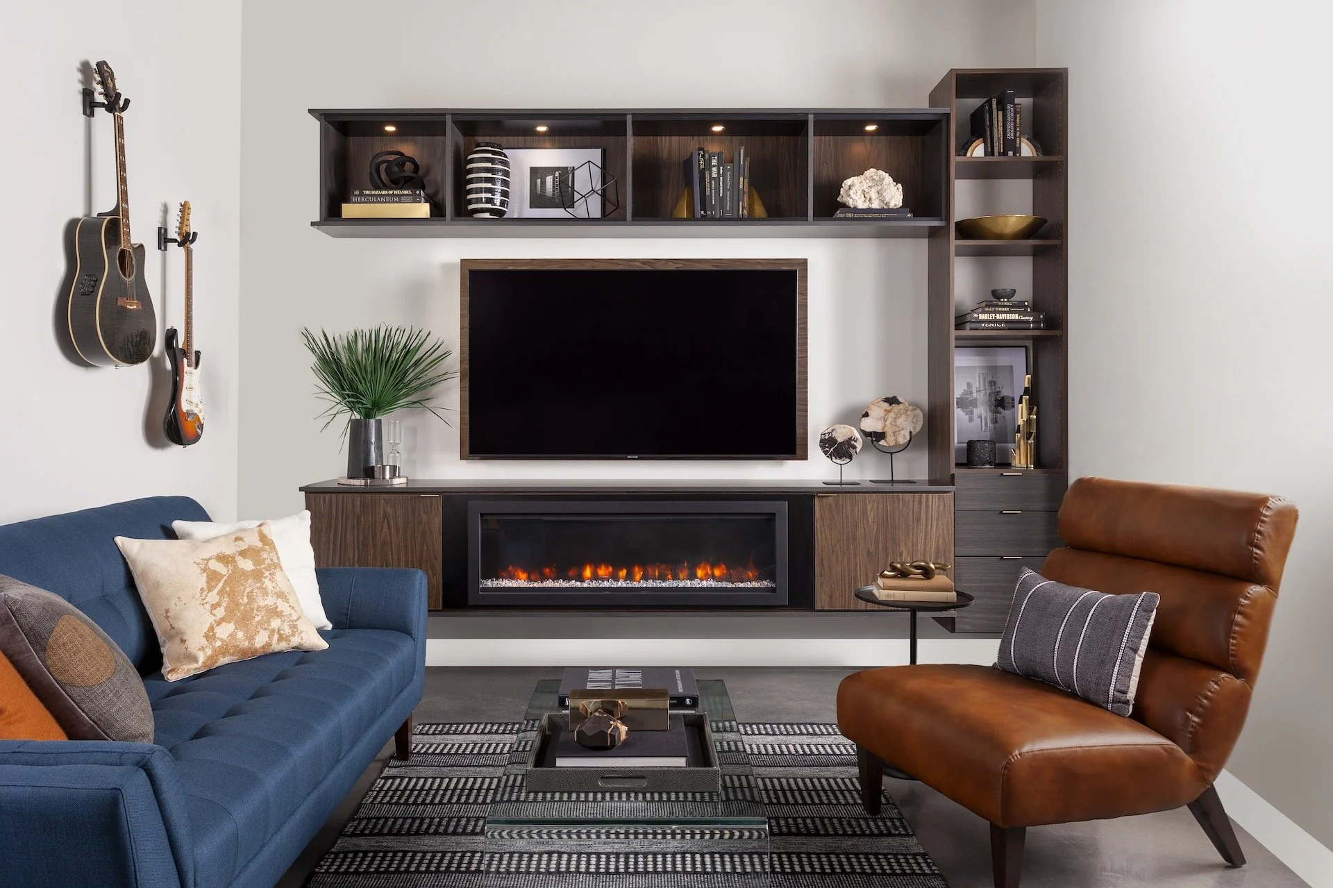Living room with wall-mounted guitars, modern wooden entertainment unit with a flat-screen TV and electric fireplace console, blue sofa with decorative pillows, brown leather chair, coffee table with books and decor, striped rug.