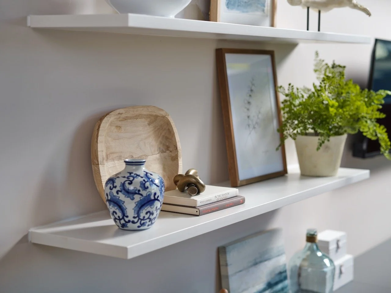 White wall-mounted shelves with decorative items including a blue and white porcelain vase, a wooden tray, a framed botanical print, a potted fern, and books.