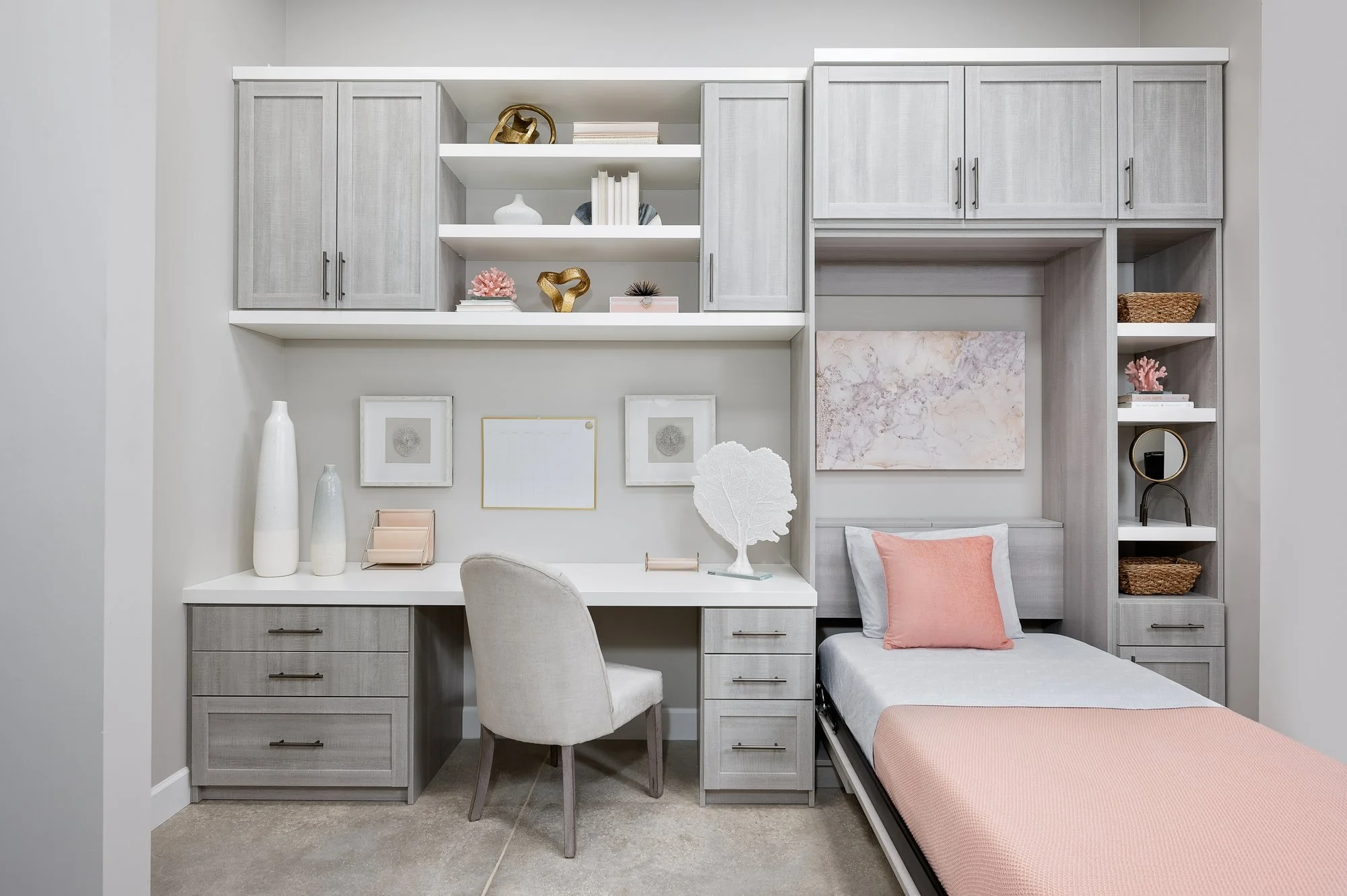 A bedroom featuring a built-in gray wooden desk and cabinets with several drawers, a white upholstered chair, and an open murphy bed with pink and white bedding.