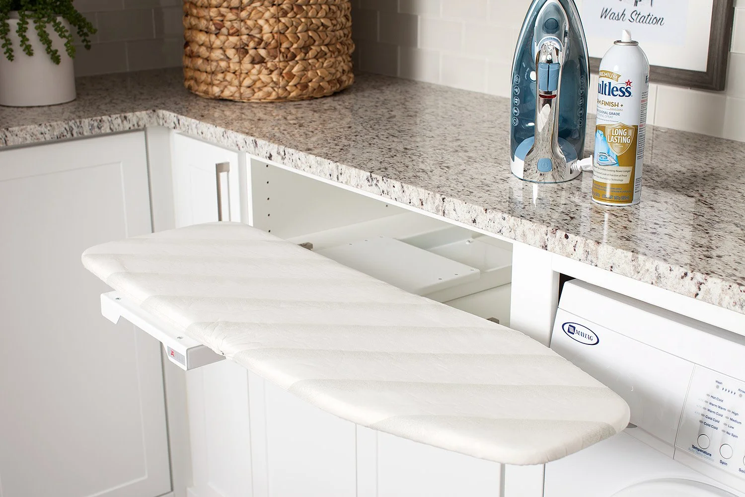 An ironing board with a white cover extended out from a cabinet drawer in front of a laundry machine in a kitchen, with a granite countertop, a soap dispenser, and an iron on the countertop.