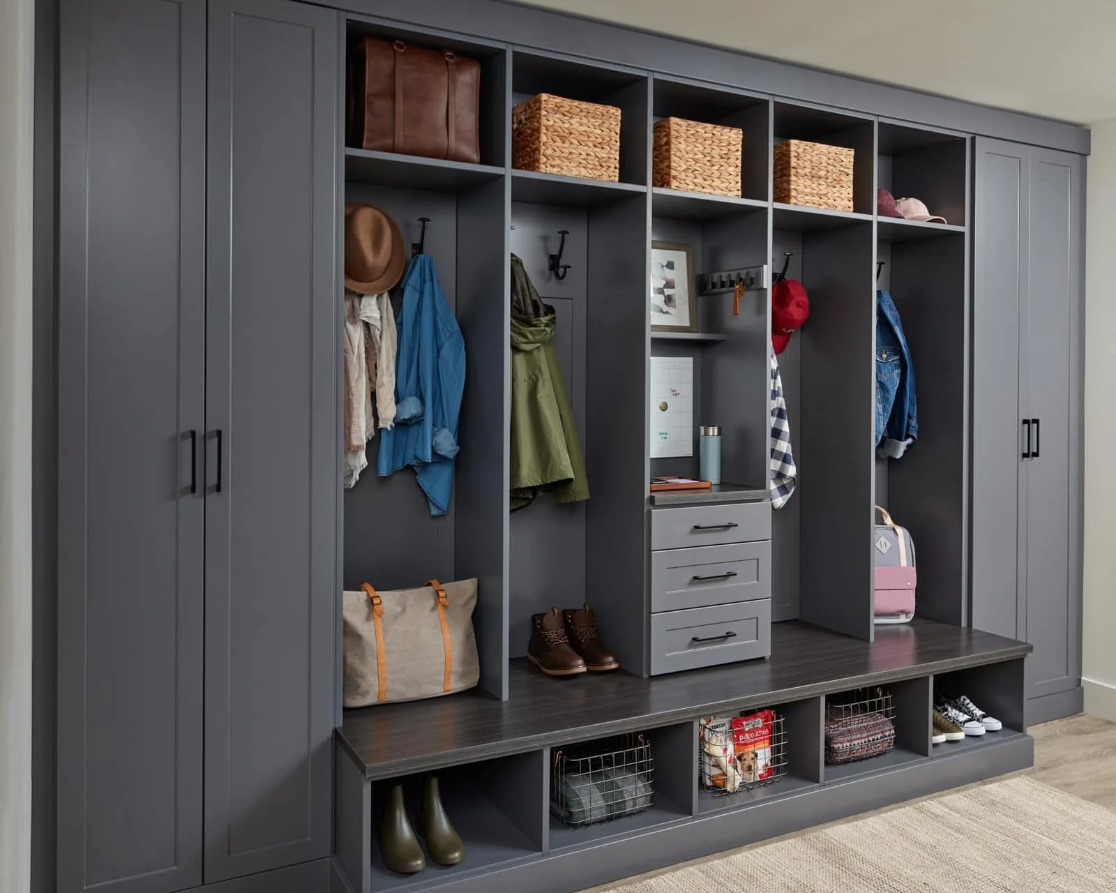 A modern, dark gray built-in entryway storage unit with hanging hooks, shelves, drawers, and cubby spaces filled with coats, hats, bags, shoes, and decorative baskets.