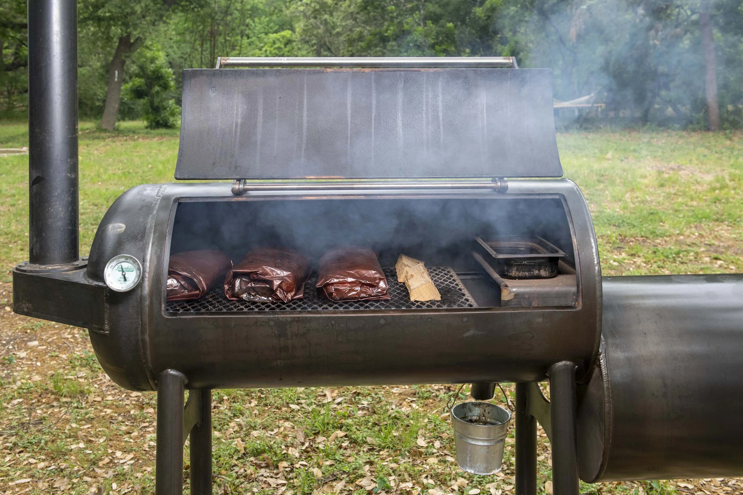 A black Franklin smoker grilling outdoors with four wrapped packages of meat on the grill and a piece of smoking wood, surrounded by a grassy area with trees.