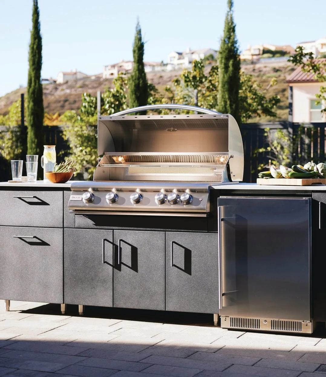 Outdoor stainless steel grill on a black counter with kitchen items and a garden in the background.