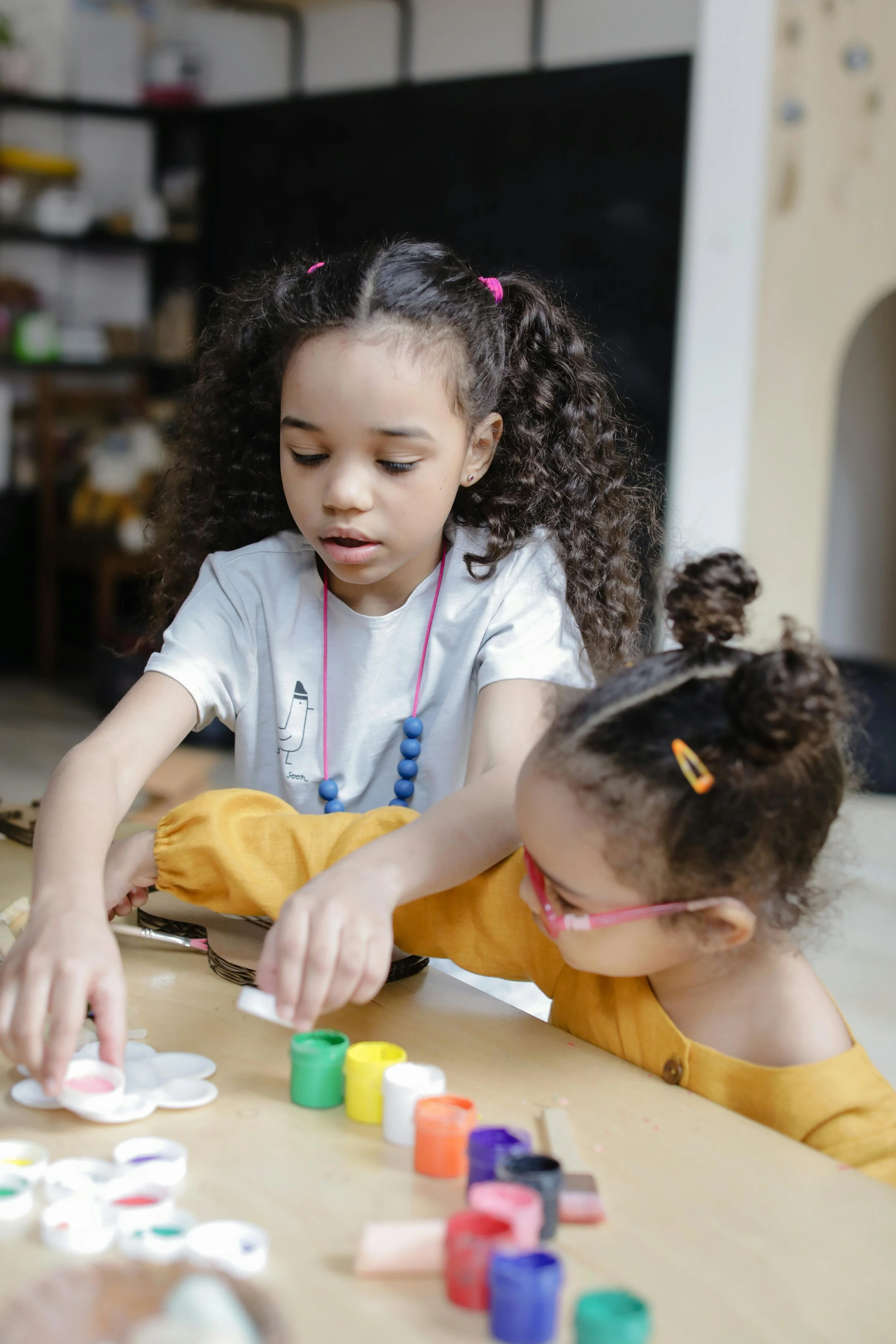 Two young girls with curly hair are playing with colourful paints and various crafts on a table.