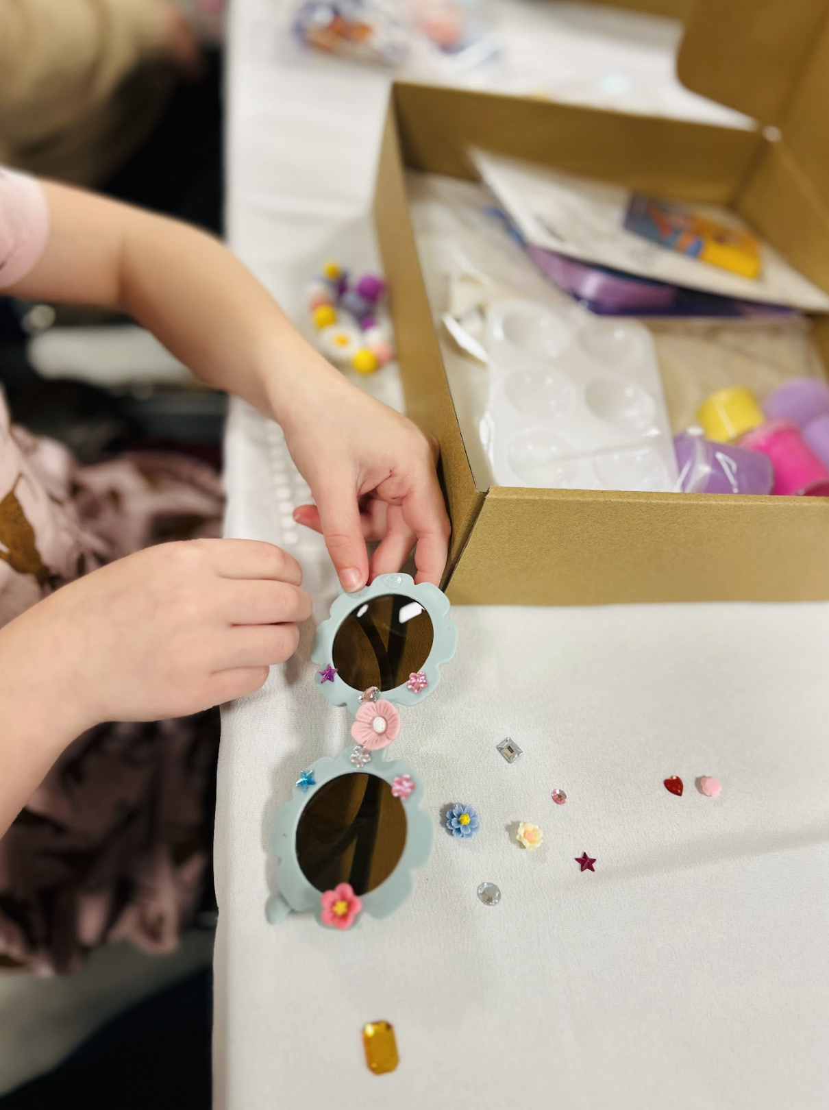 Child decorating a pair of sunglasses with floral and star-shaped embellishments, next to a box of craft supplies and scattered decorative stickers and gems on a white table.