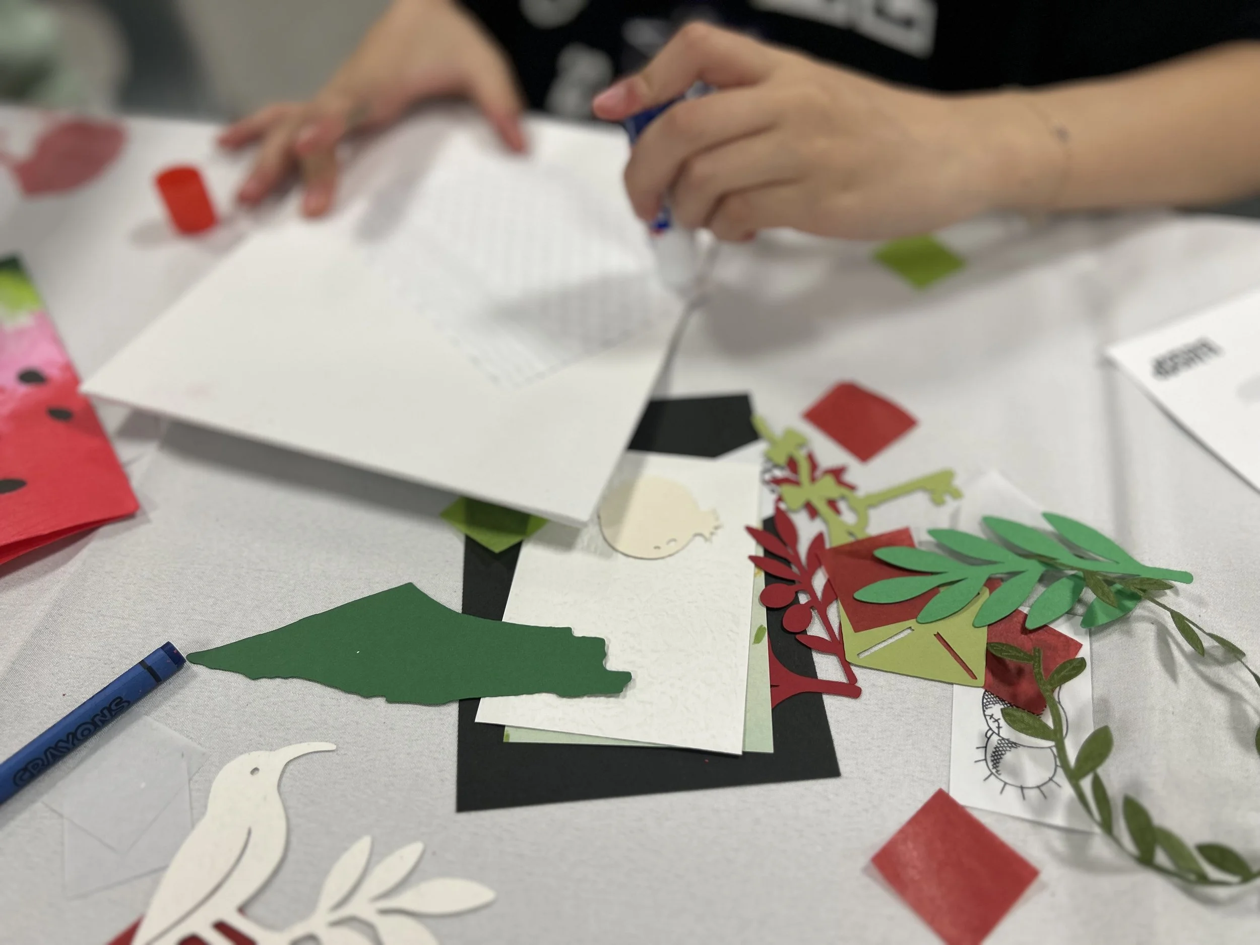 Person making a holiday craft with paper cutouts of leaves and other shapes, sitting at a table covered with holiday-themed paper and decorations.