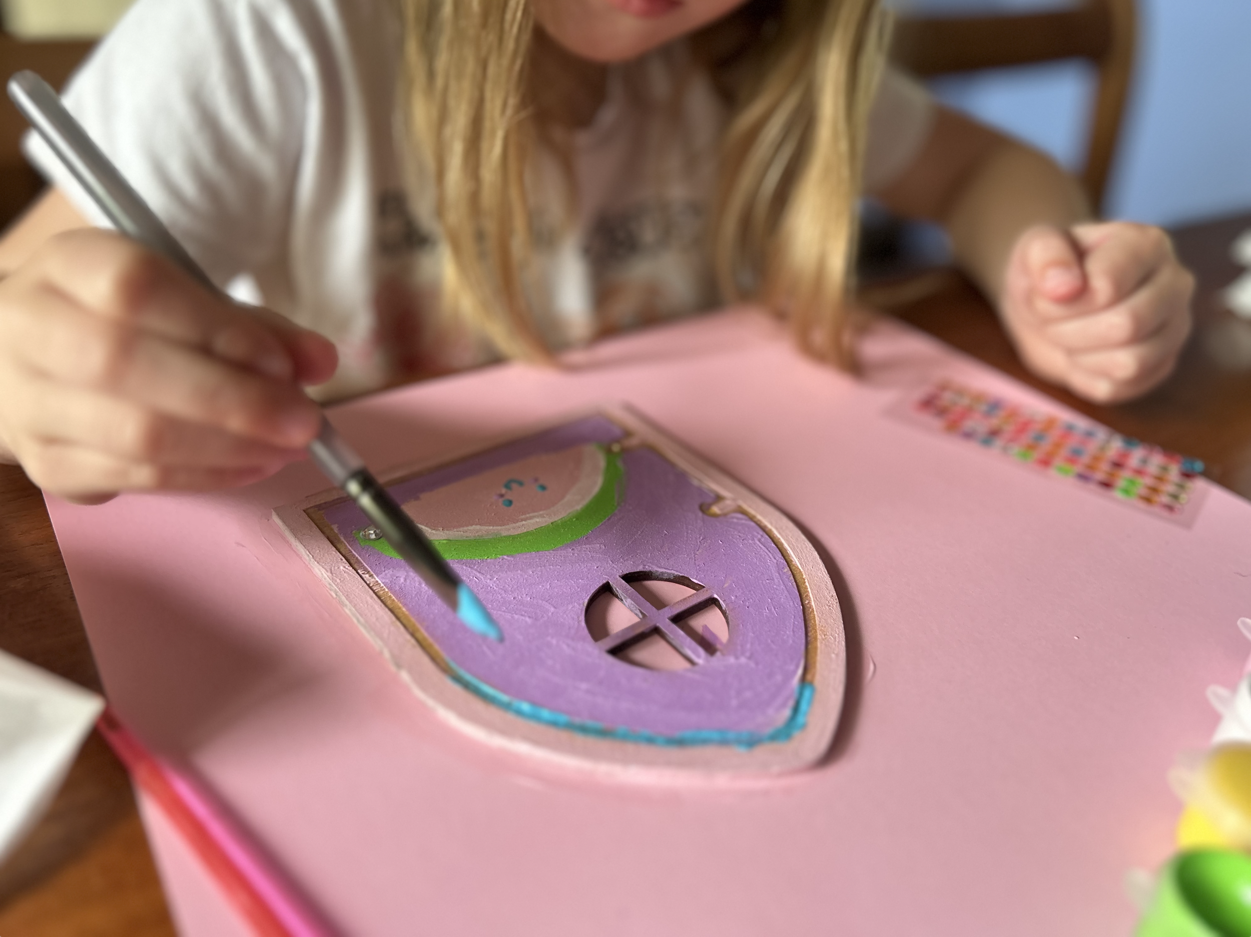 A young girl decorates a heart-shaped wooden craft with colorful paint, sitting at a pink table.