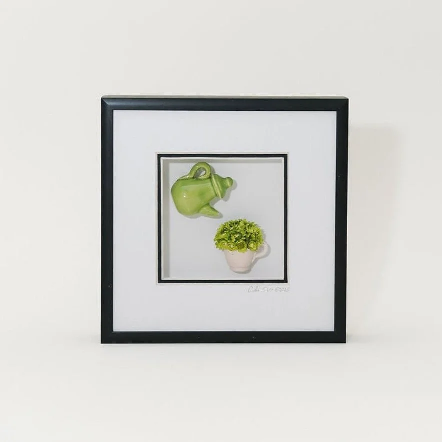 Framed artwork featuring a green teapot and a small white cup with green dried flowers.