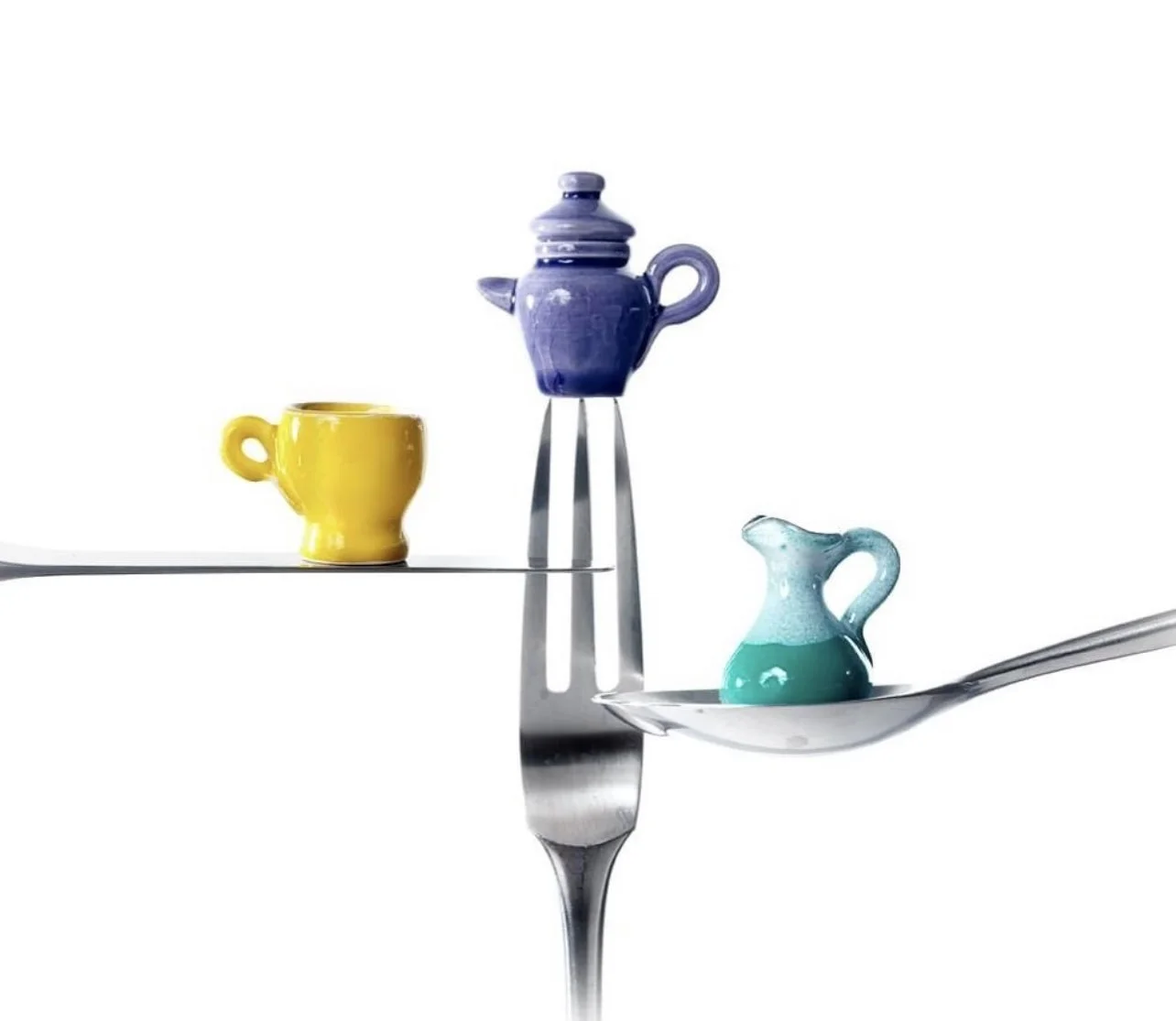 Colorful ceramic teapots and cups balanced on the prongs of a large metallic fork and a knife against a plain white background.