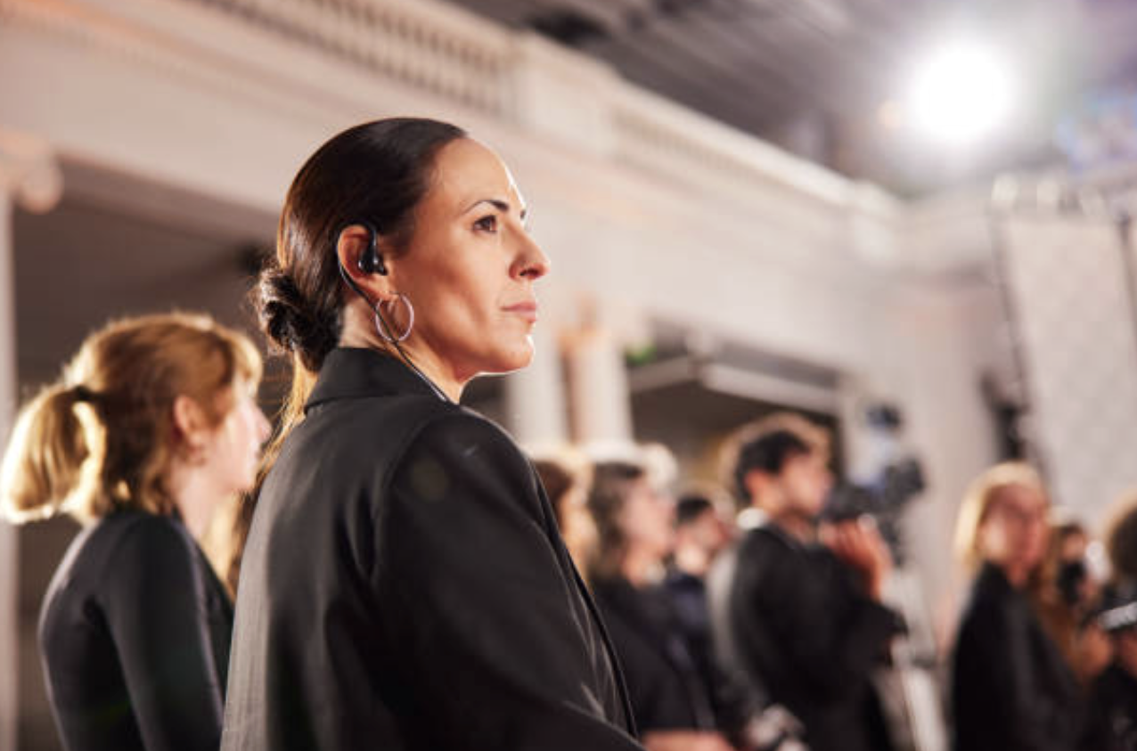 A woman with dark hair styled in a bun, wearing a black blazer and an earpiece, attentively watching an event in a large indoor setting with other people in the background.