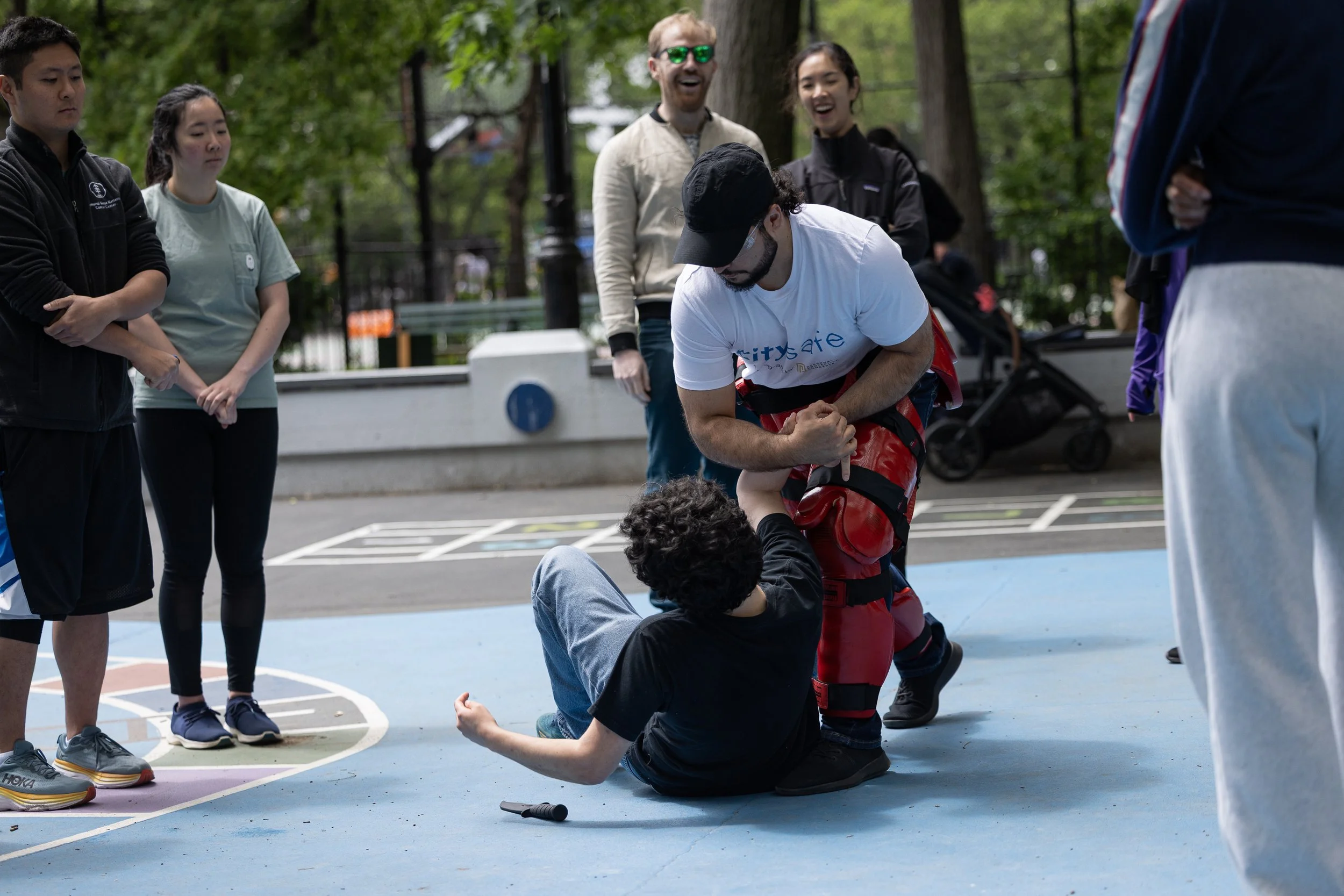 A man in a white shirt and red protective gear is helping another man to the ground during a self-defense or martial arts demonstration on an outdoor basketball court, while several people watch.