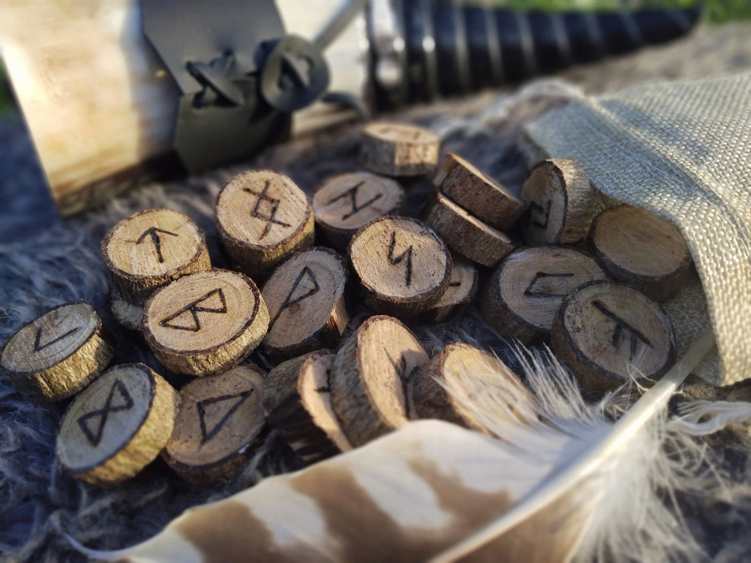 Small wooden discs with runes inscribed, scattered on a textured surface, with a feather and a cloth nearby, and a carved wooden object in the background.