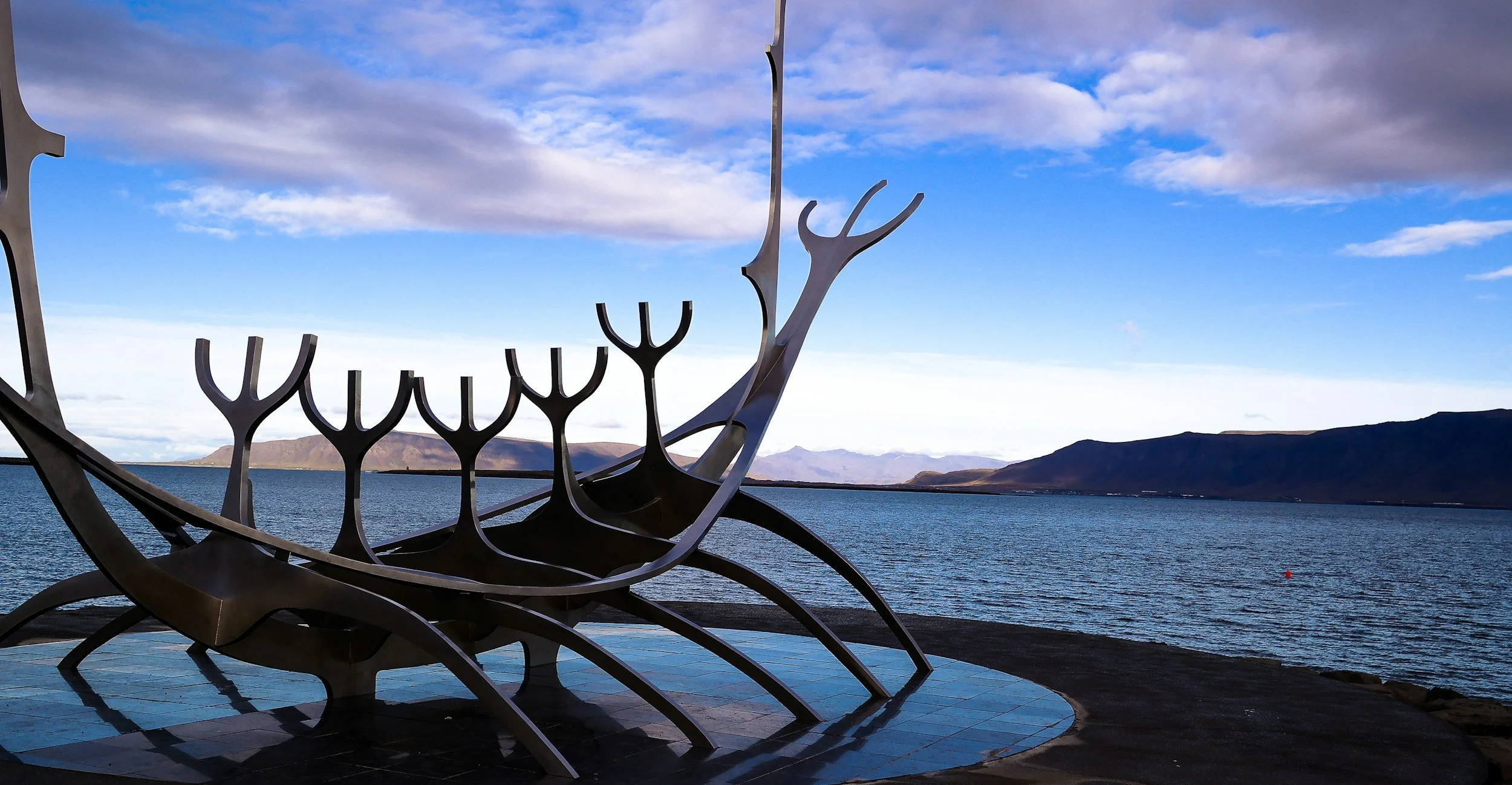 A metallic sculpture of a Viking ship with a dragon-shaped prow on the shore, with mountains across a body of water under a blue sky with clouds.