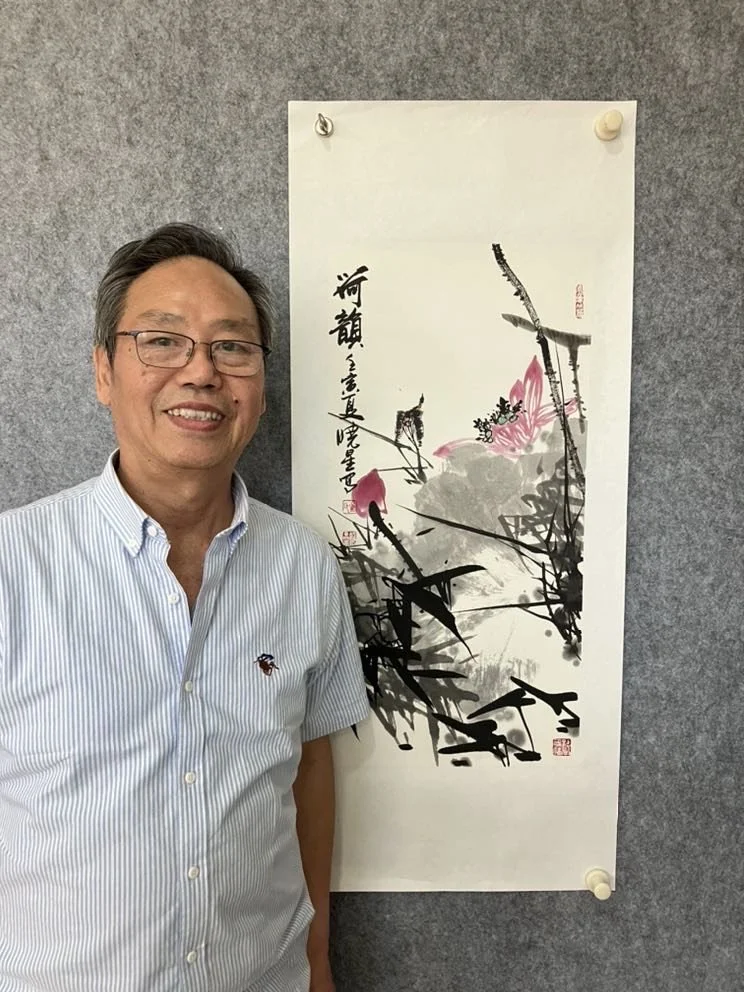 A smiling man standing next to a traditional Chinese ink painting on a scroll, featuring pink flowers and black brushstrokes, mounted on a gray wall.