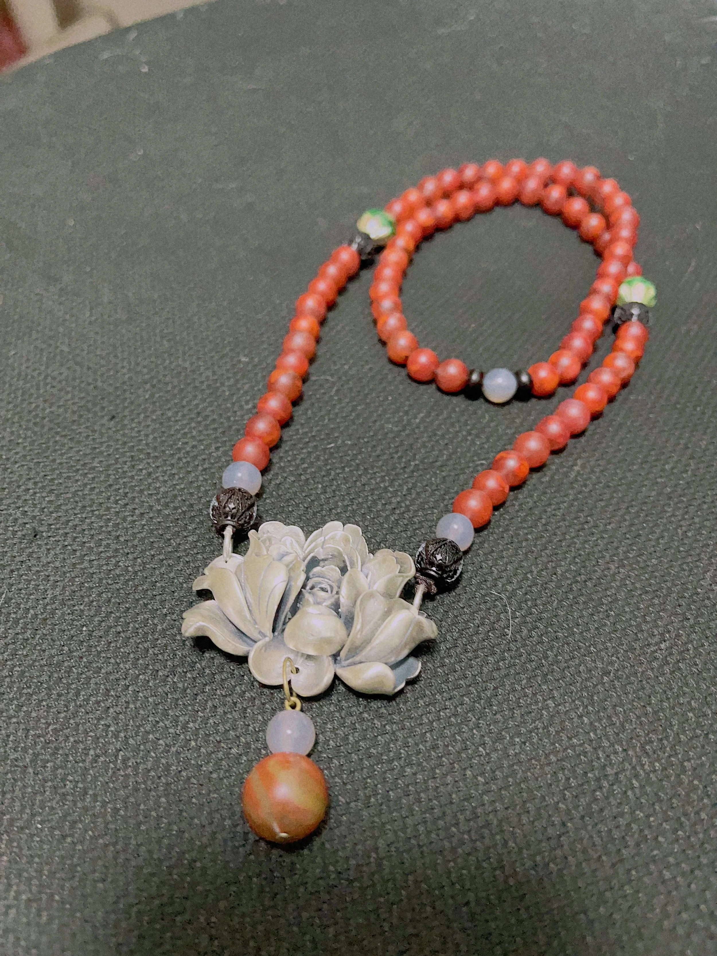 A beaded necklace with orange, gray, and black beads, featuring a large carved floral pendant with a dangling orange bead.