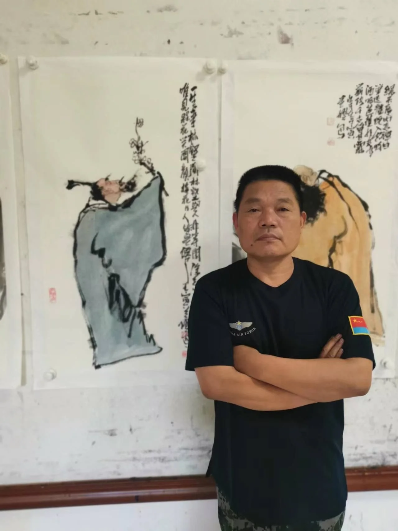 A man with black hair, wearing a black T-shirt with military patches, stands with arms crossed in front of traditional Chinese calligraphy paintings on the wall.