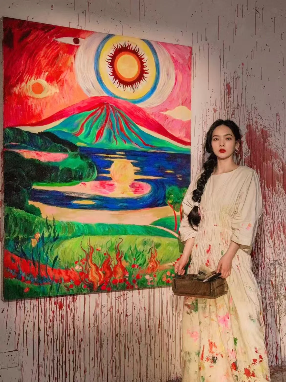 A young woman with long dark hair in a braid, wearing a cream-colored dress with paint splatters, stands in front of a colorful painting of a mountain landscape with a large sun and a river. The wall behind her is splattered with red paint or blood.