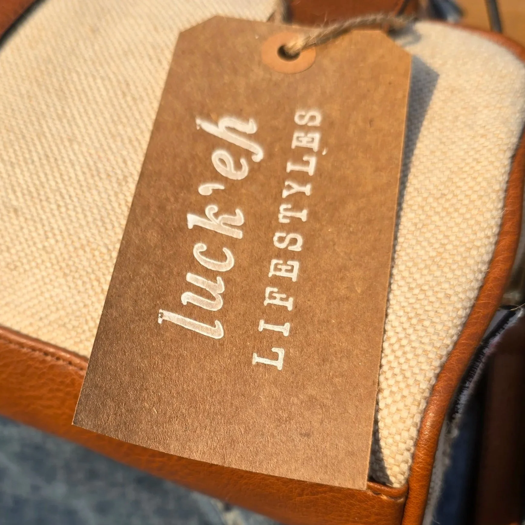 Close-up of a brown craft paper tag with a brand label stamped that reads 'luck'eh lifestyles'. The tag is placed on a beige textured surface (bag).