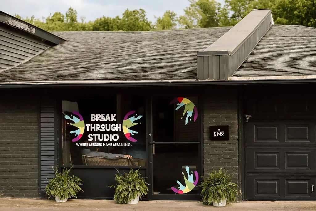 Exterior of a black brick building with a sign that says "Break Through Studio" and the slogan "Where messes have meaning." Potted plants are in front, and colorful hand and paint splash graphics decorate the windows and door.