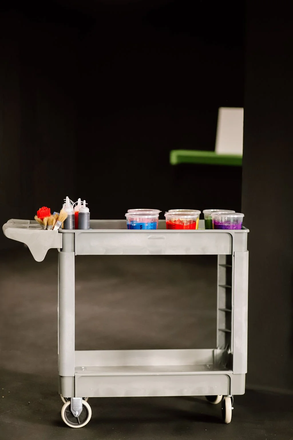 A metal cart with various supplies including paint bottles, brushes, and cups of colorful liquid in front of a black wall.