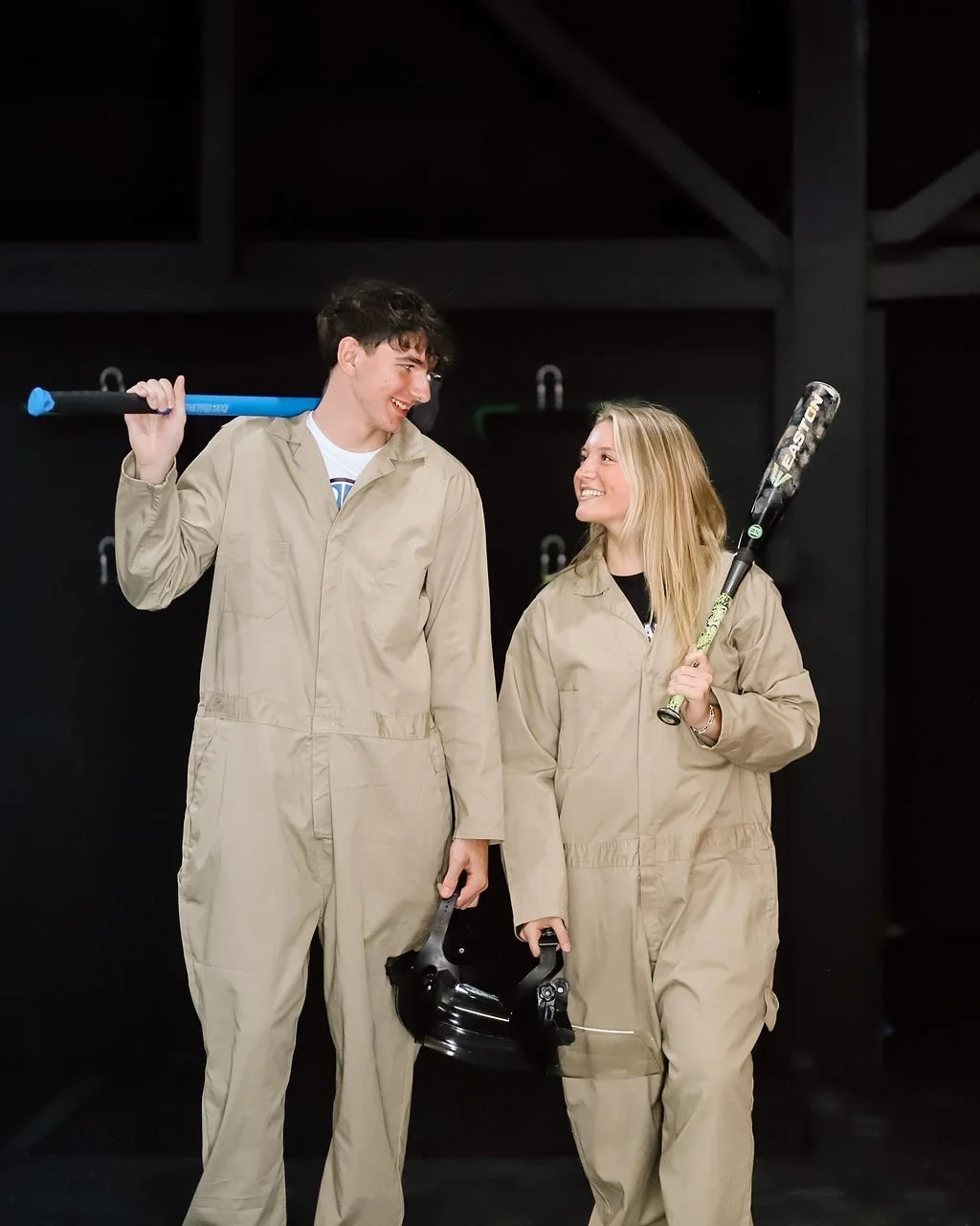 A young man and woman in jumpsuits smiling at each other while holding baseball bats.