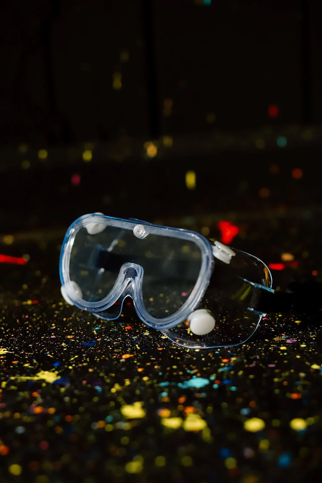 A pair of clear safety goggles resting on a black surface covered in multicolored glitter, with a blurred cityscape at night in the background.