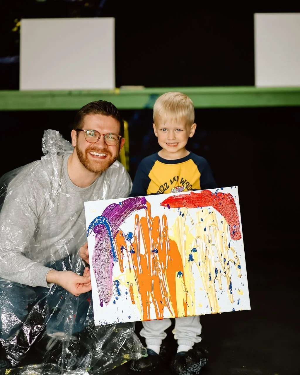 A man and a young boy smiling and holding a colorful abstract painting together. The man is wearing glasses and a silver raincoat, and the boy has blond hair and is wearing a blue and yellow shirt with white pants.