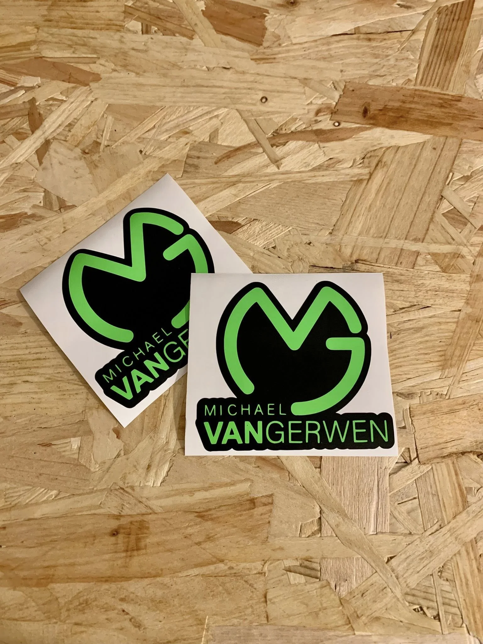 Two stickers with a black and green logo and the name 'Michael Vangerwen' on a wood panel background.