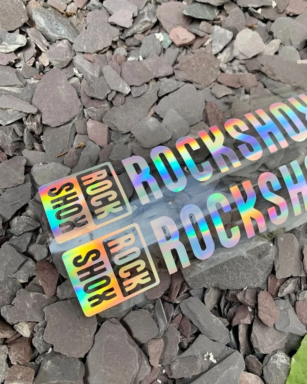 Holographic stickers with the text 'BOOK STORE ROCK SHOP' placed on a bed of small, dark rocks.