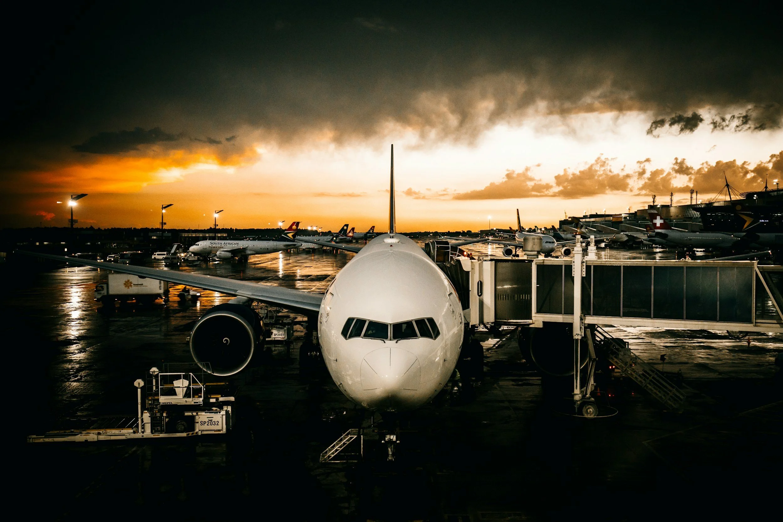 View of an airport tarmac at sunset with multiple airplanes parked at gates, one airplane in the foreground, and a dramatic dark sky with clouds and orange glow.