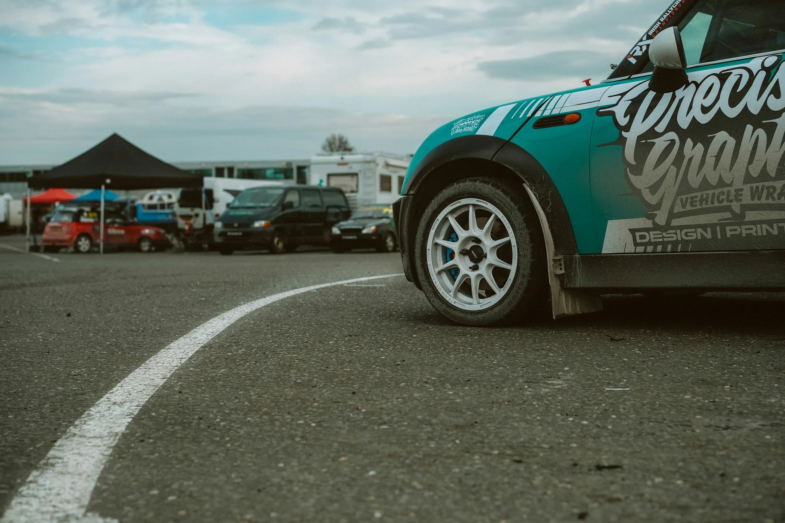 Close-up of a teal race car with large decal designs on the side, parked on an asphalt lot with other cars and tents in the background.