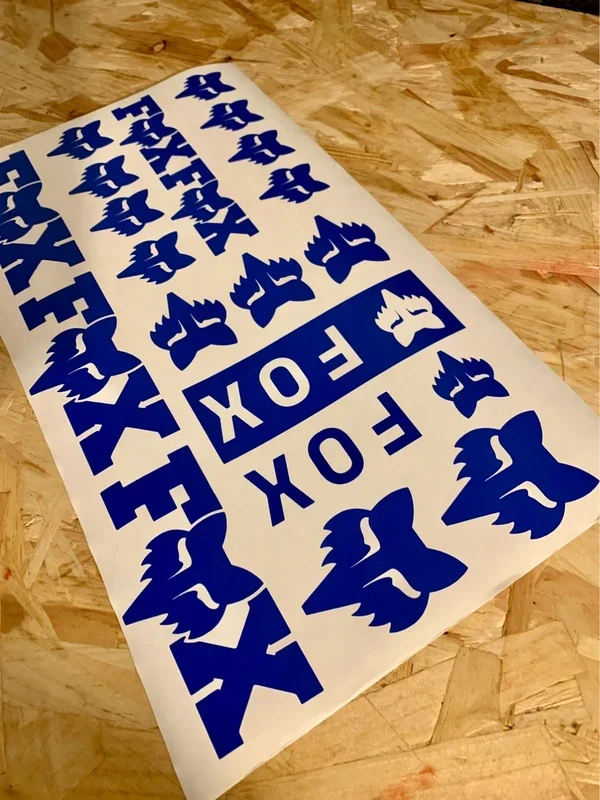 Sheet of blue and white stickers with Fox logo and text reading "FOX" and "FOX" repeated, some featuring a fox head graphic, placed on a wooden surface.